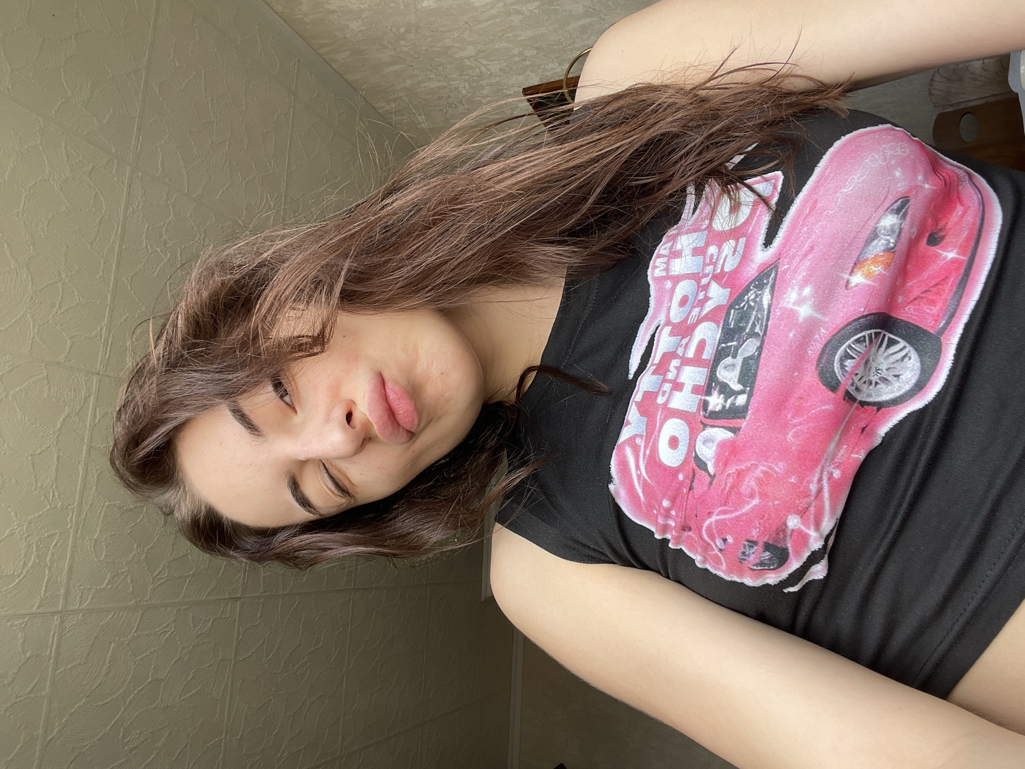 A young woman with long brown hair is lying down and facing the camera. She is wearing a black t-shirt with a pink car graphic. The background is a textured wall. The image appears to be taken indoors. The location in Moscow, Russia is not evident from the image.