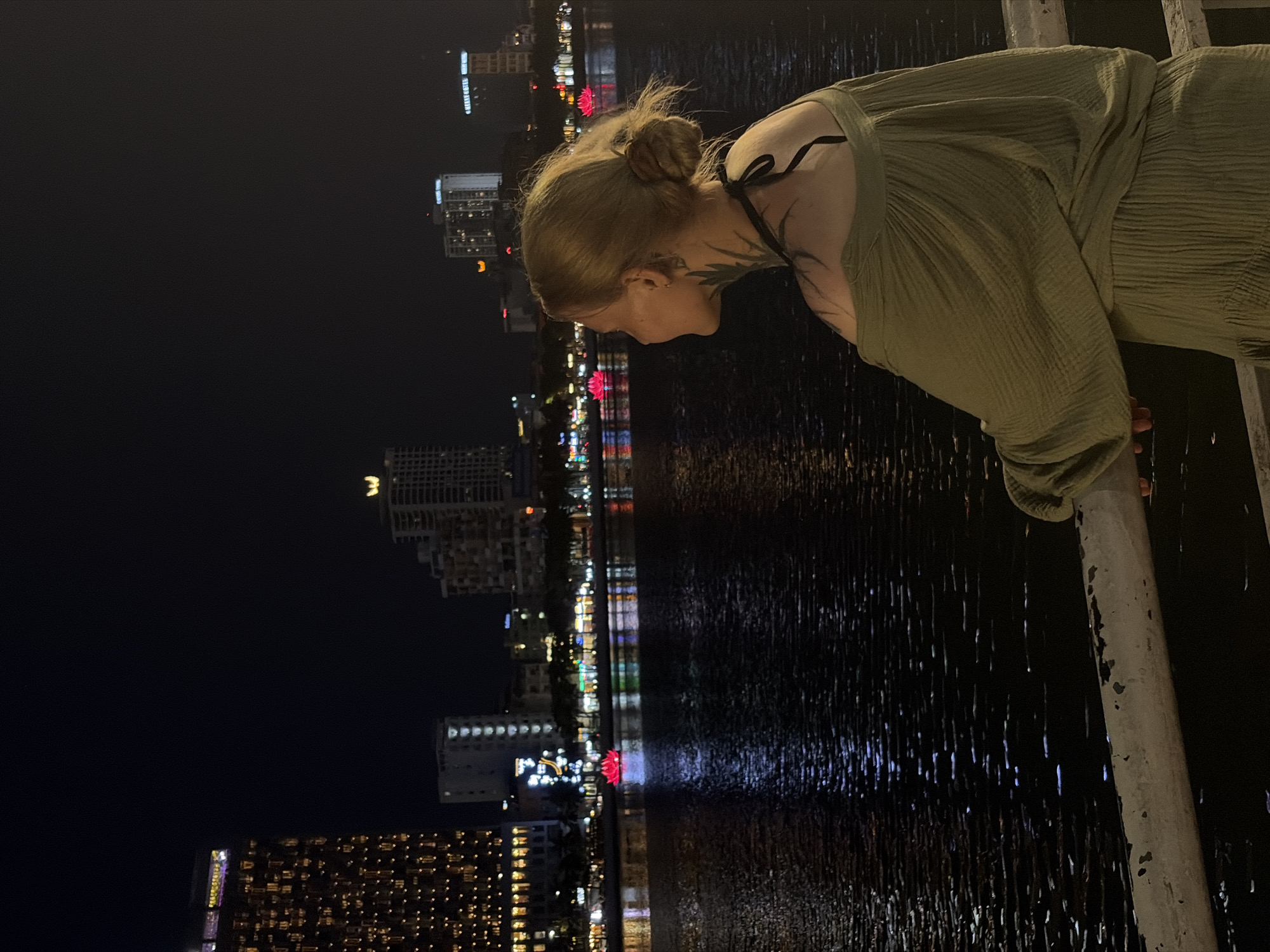 The image depicts a nighttime scene in Đà Nẵng, Vietnam, featuring a young woman leaning over a weathered white railing, gazing at the illuminated city reflected in the dark water. The woman is shown in profile from the waist up, with light-colored hair pulled back into a bun. She wears a loose-fitting, textured olive-green or khaki top, and intricate dark, leafy tattoos are visible on her neck and extending towards her shoulder, accompanied by a thin black strap or ribbon. Her hands are placed gently on the railing as she looks downward, suggesting a moment of quiet contemplation or observation.
Across the body of water, which is likely a river or bay, a vibrant city skyline is visible, characterized by numerous brightly lit high-rise buildings. Their lights create sparkling, elongated reflections on the water's surface, contributing to the serene nocturnal ambiance. A distinct red, lotus-shaped light is prominent among the reflections or on the distant shore.
The scene conveys a peaceful atmosphere, with the city lights providing the primary illumination. Visible text on some of the distant buildings includes "NOVOTEL" and "GOLDEN BAY". The railing she leans on shows signs of wear, with peeling paint.