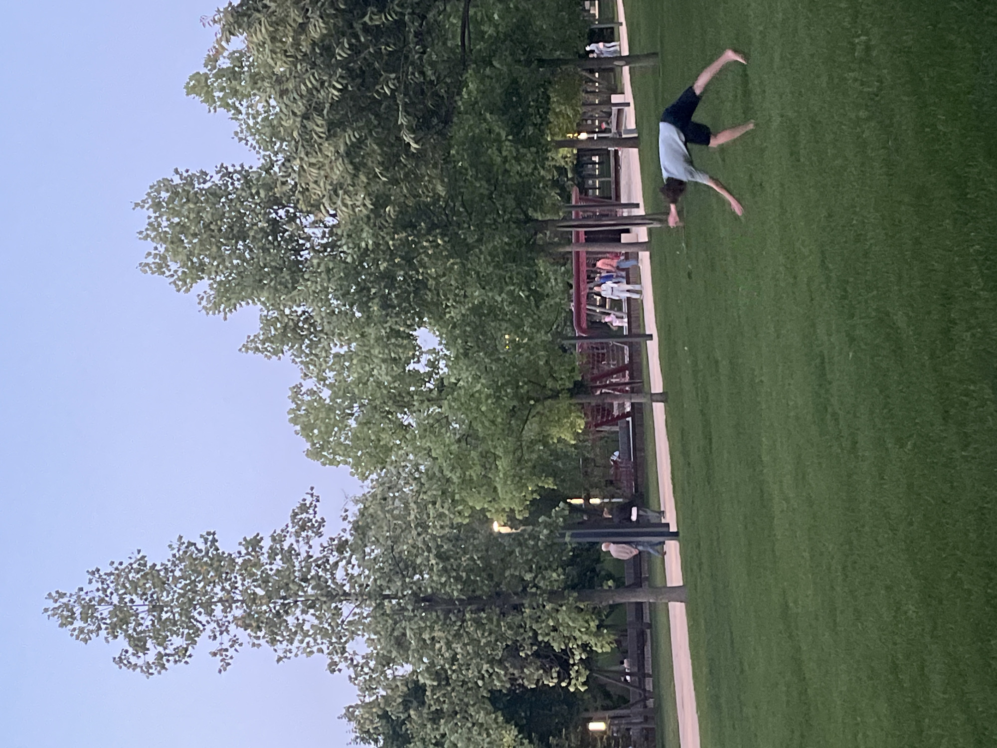 The image shows a person performing a cartwheel on a grassy area, likely a park. The individual is wearing a light-colored shirt and dark shorts. In the background, a tree line separates the grassy area from a pedestrian walkway, with other people and some structures visible further back. The sky is a pale blue, suggesting either dawn or dusk. The location in the city of Krasnodar, Russia cannot be verified from visual data.