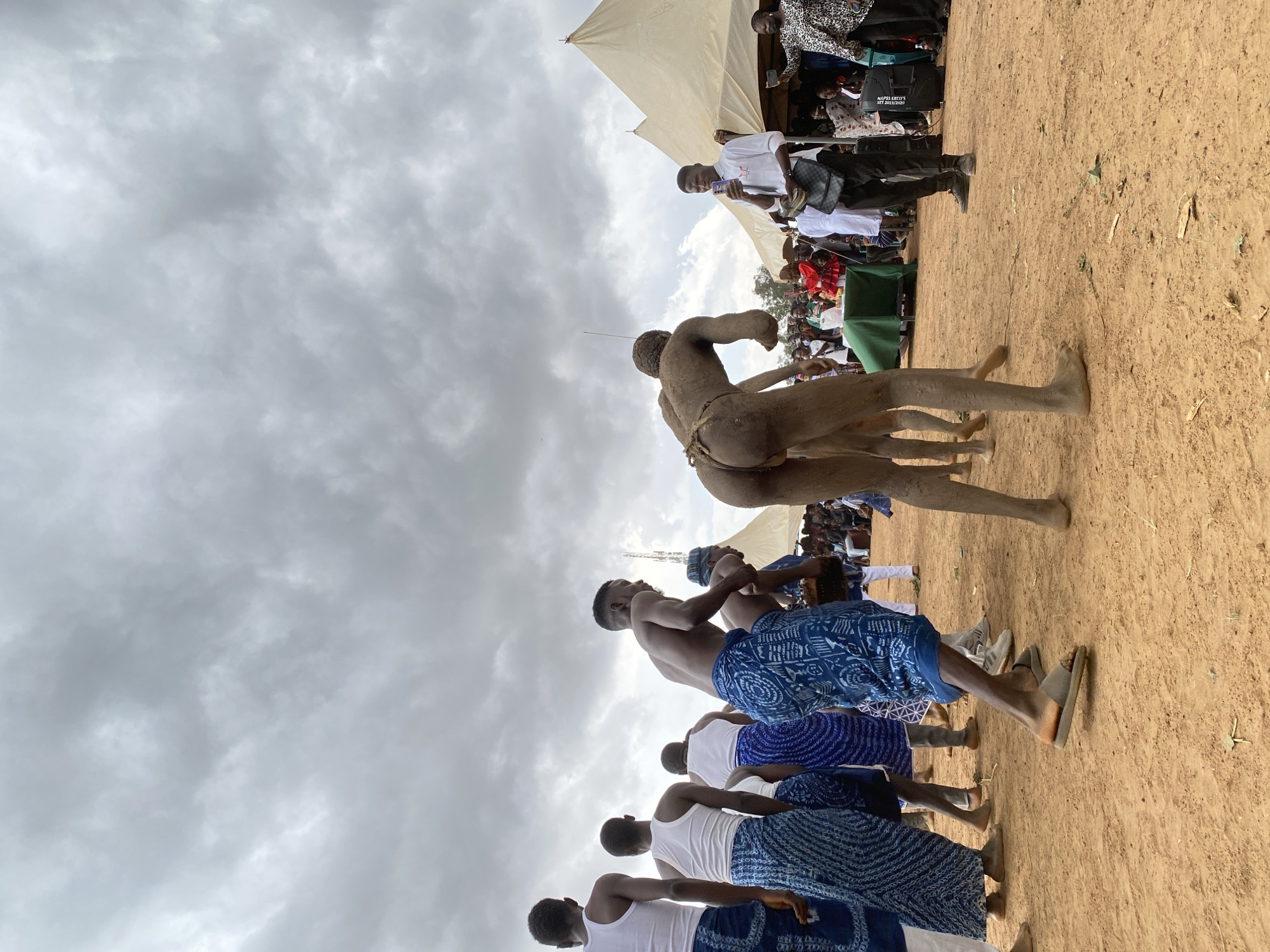 The image depicts a traditional wrestling match taking place outdoors on a dirt surface in the city of Durimi, Nigeria. Two nude wrestlers are the focal point, covered in dirt, locked in a grappling hold. A group of men, some wearing white tops and blue patterned wraps around their waists, are positioned near the wrestlers. Spectators are seen in the background under a tent-like structure. The sky is overcast with heavy clouds.