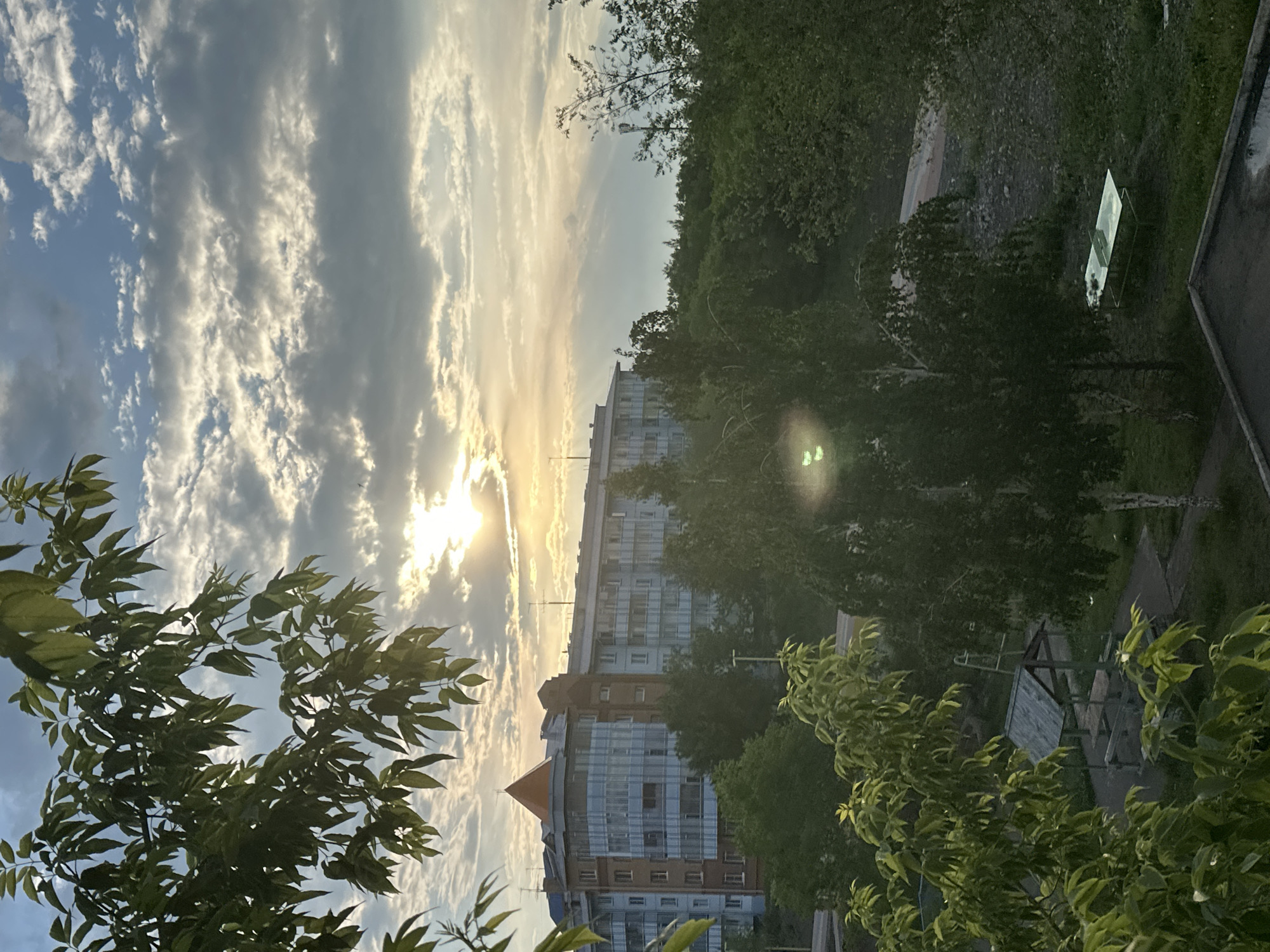The image captures a late afternoon or early evening scene in Sayanogorsk, Russia, characterized by soft, diffused sunlight breaking through a sky filled with scattered clouds. In the foreground, leafy branches of deciduous trees frame the view. Dominating the mid-ground is a multi-story residential building with a distinctive orange-capped peak. The building's facade features a mix of brick and glass sections, with balconies visible. Beyond the building, dense green trees form a natural backdrop. In the lower part of the image, there are glimpses of outdoor recreational amenities, including a covered picnic area with benches and a ping pong table. The overall atmosphere is tranquil, with the natural light creating a serene, almost pastoral, feel despite the urban elements.