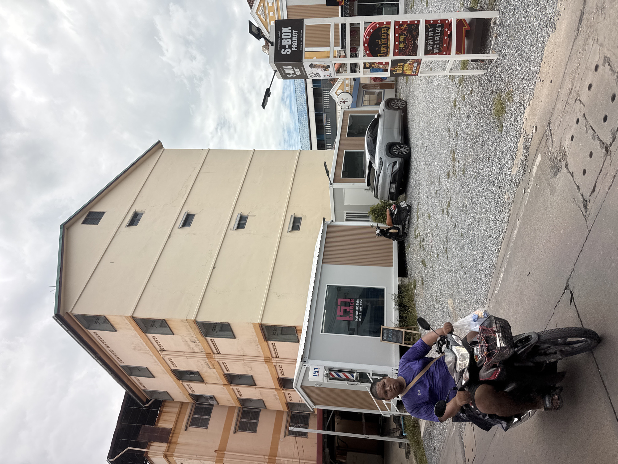 A person rides a motorcycle on a paved road in front of a building complex. The person is wearing a purple shirt and black shorts, and is riding a black motorcycle with a basket in the front. The building complex has several floors and is painted in light yellow and orange colors. In front of the building, there are several small, modern modular buildings, including a barber shop with a red and white striped pole. A silver electric car is parked nearby. A signpost with the text "S-BOX PROJECT" is visible. The ground is covered with gravel. The sky is cloudy. This scene is in Nonthaburi City Municipality, Thailand.