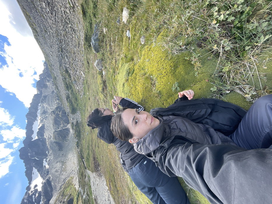 Two women lie on their backs on mossy ground. The woman in the foreground has short brown hair, a light-colored vest, and dark pants. The other woman, behind her, has dark hair and wears an Adidas tracksuit. A large, rocky mountain rises steeply to the left, with patches of snow. The sky is visible above, with some clouds. The ground appears to be a hillside.