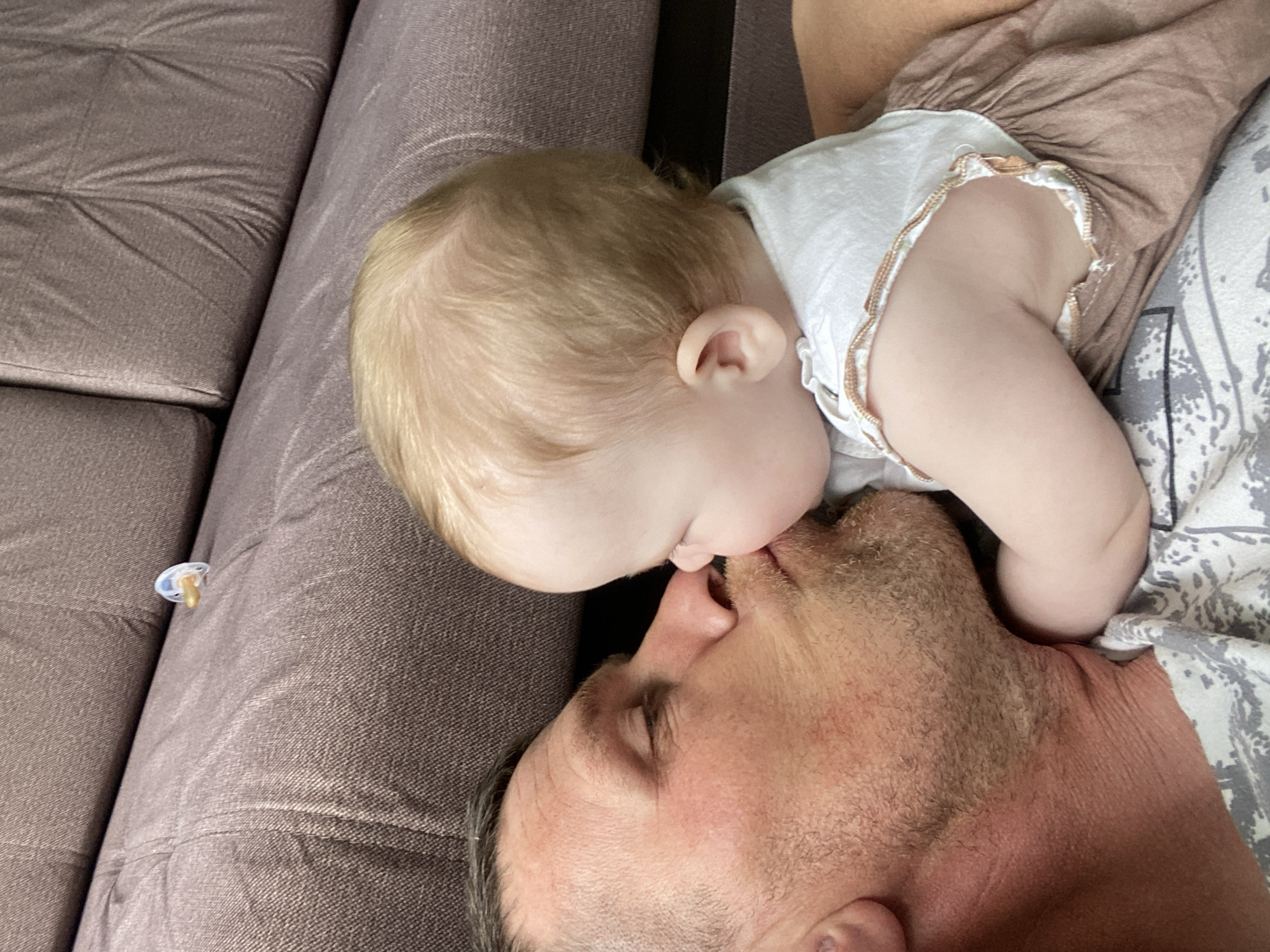 A medium shot shows a Caucasian man lying on his back, eyes closed, on a brown couch. A fair-skinned infant with light blonde hair is lying on the man's chest, its lips touching his nose. The baby is wearing a white and brown ruffled garment. A pacifier is visible on the couch near the man's head. The man is lying on a patterned gray and white fabric. The scene does not seem to be in the city of Фоки, Russia.