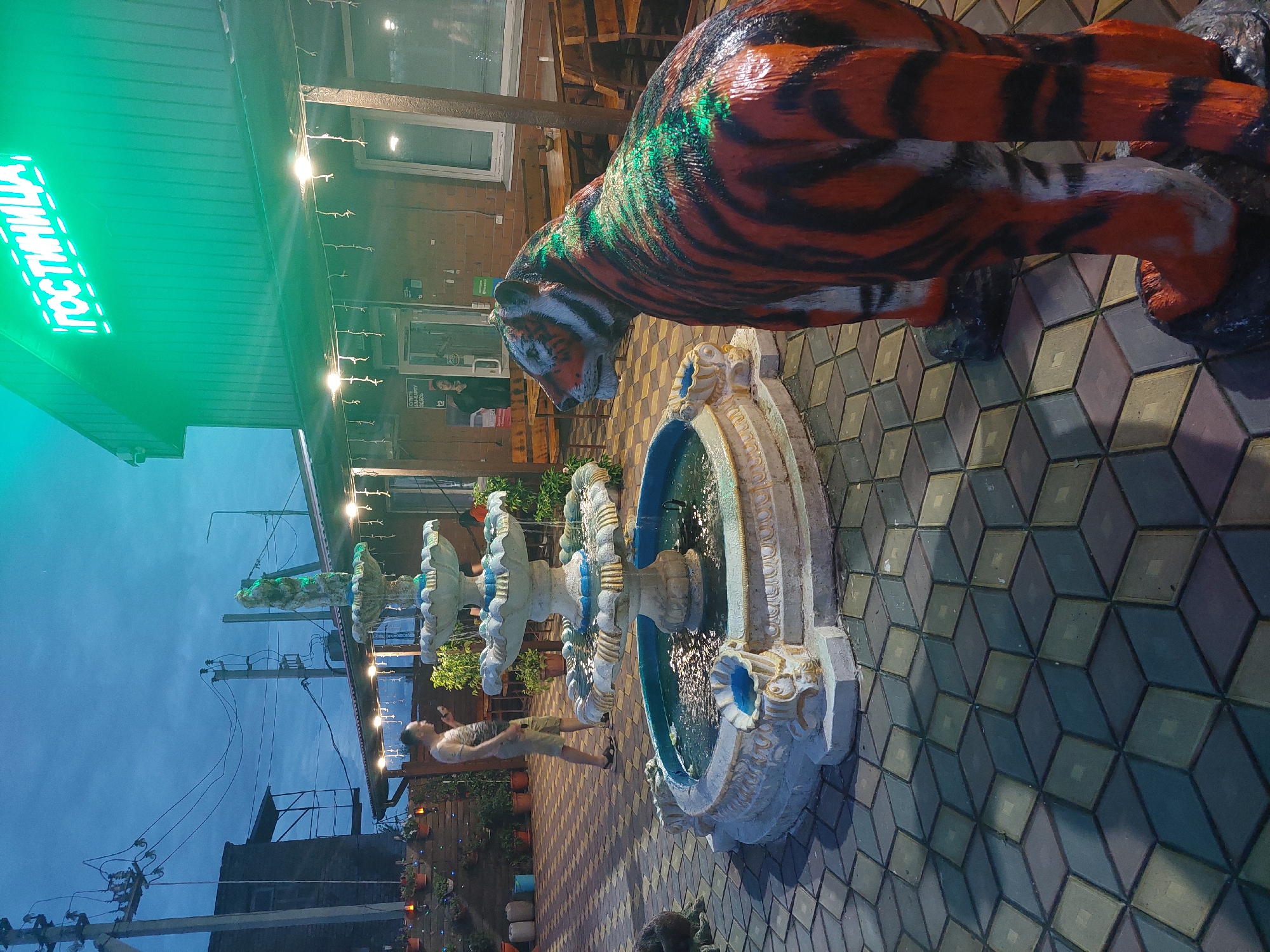 The image depicts an outdoor seating area adjacent to a building with a neon green sign that reads "ГОСТИНИЦА" (HOTEL). In the foreground, a statue of a tiger, painted orange and black with green accents, stands on a base. To the left, a multi-tiered fountain with blue trim and a sculpture of a figure at its apex is active. A man in a light-colored t-shirt and shorts is standing near the fountain, holding a phone. The ground is paved with interlocking geometric tiles. In the background, tables and benches suggest an outdoor dining or seating area. The sky is dark, indicating dusk or evening. The overall scene appears to be an entrance or courtyard of a hotel in Dalneretschensk, Russia.