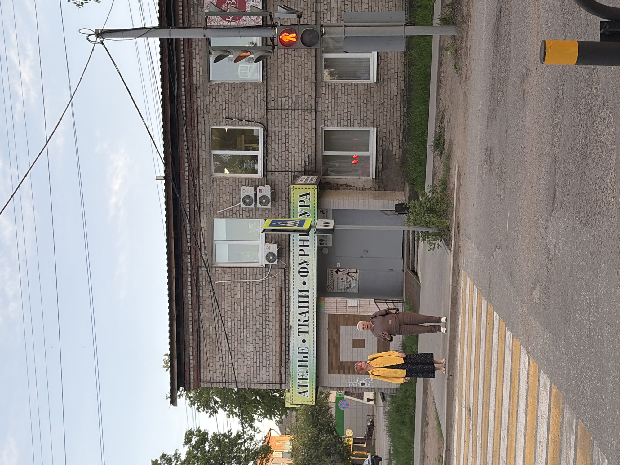 The image depicts a street scene in what appears to be the city of Chaikovsky, Russia. Two individuals are crossing a crosswalk in front of a two-story brick building. The woman on the right has a yellow jacket, the woman on the left is wearing a brown jumpsuit and carrying a bag. Above the crosswalk, there is a building. It has several windows and two air conditioning units attached to the side. A vertical green sign is attached to the building. It reads "АТЕЛЬЕ · ТКАНИ · ФУРНИ," which translates to "Atelier · Fabrics · Furnishings." A traffic light displays a red pedestrian signal. Power lines are visible overhead. The street looks well-maintained and clean.