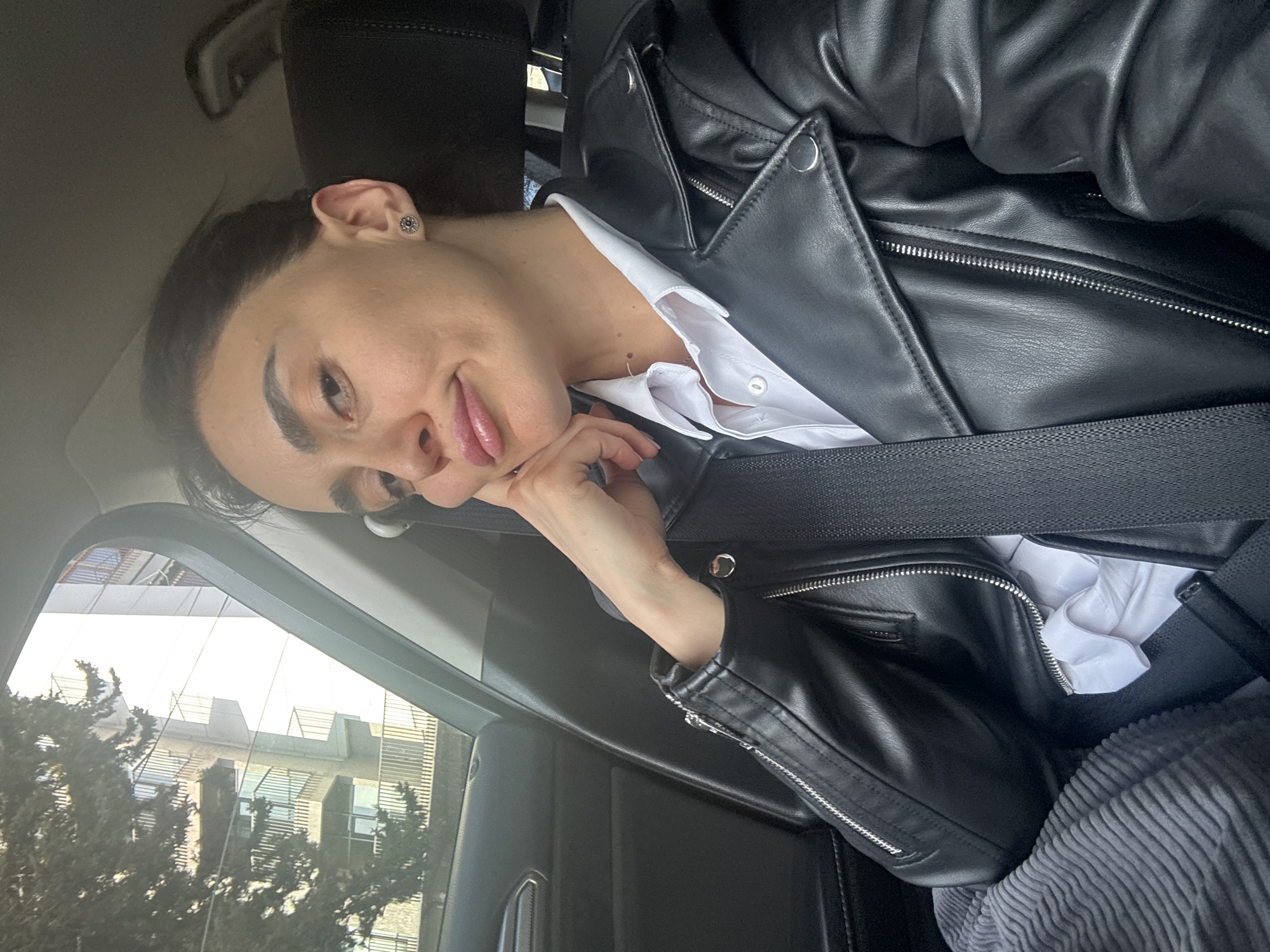 A person with dark hair is seated in a car, wearing a black leather jacket over a white collared shirt and a seatbelt. The individual's head is resting on a hand. Outside the car window, a building can be seen, suggesting the location is in the city of Tbilisi, Georgia. The interior of the car includes dark upholstery.