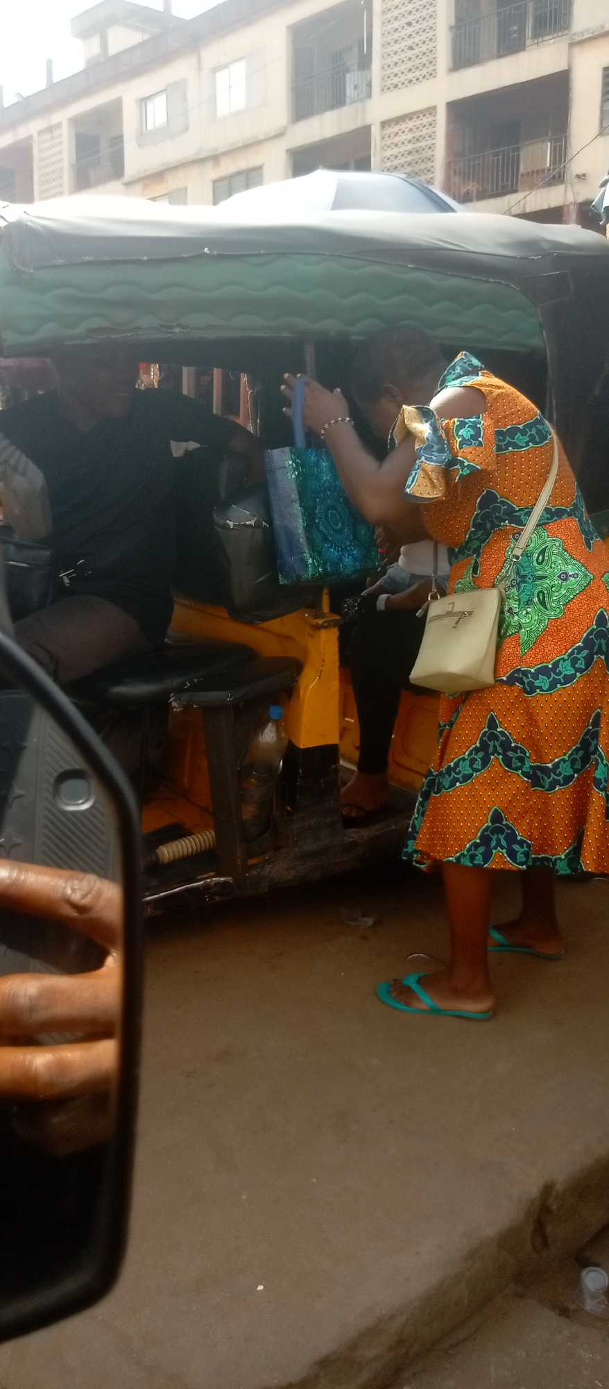 In Nkpor, Nigeria, the scene depicts a motorized tricycle (Keke Napep) at the edge of a street or road. The tricycle is yellow and black, and the driver sits inside, dressed in black, and appears to be interacting with a female passenger. The passenger stands outside the Keke Napep and is wearing an orange, green, and white patterned dress with matching blue flip-flops. She has a small white purse over her shoulder and holds a shiny blue shopping bag. The surroundings include urban buildings, and the road surface is simple, suggesting an ordinary day in Nkpor.
