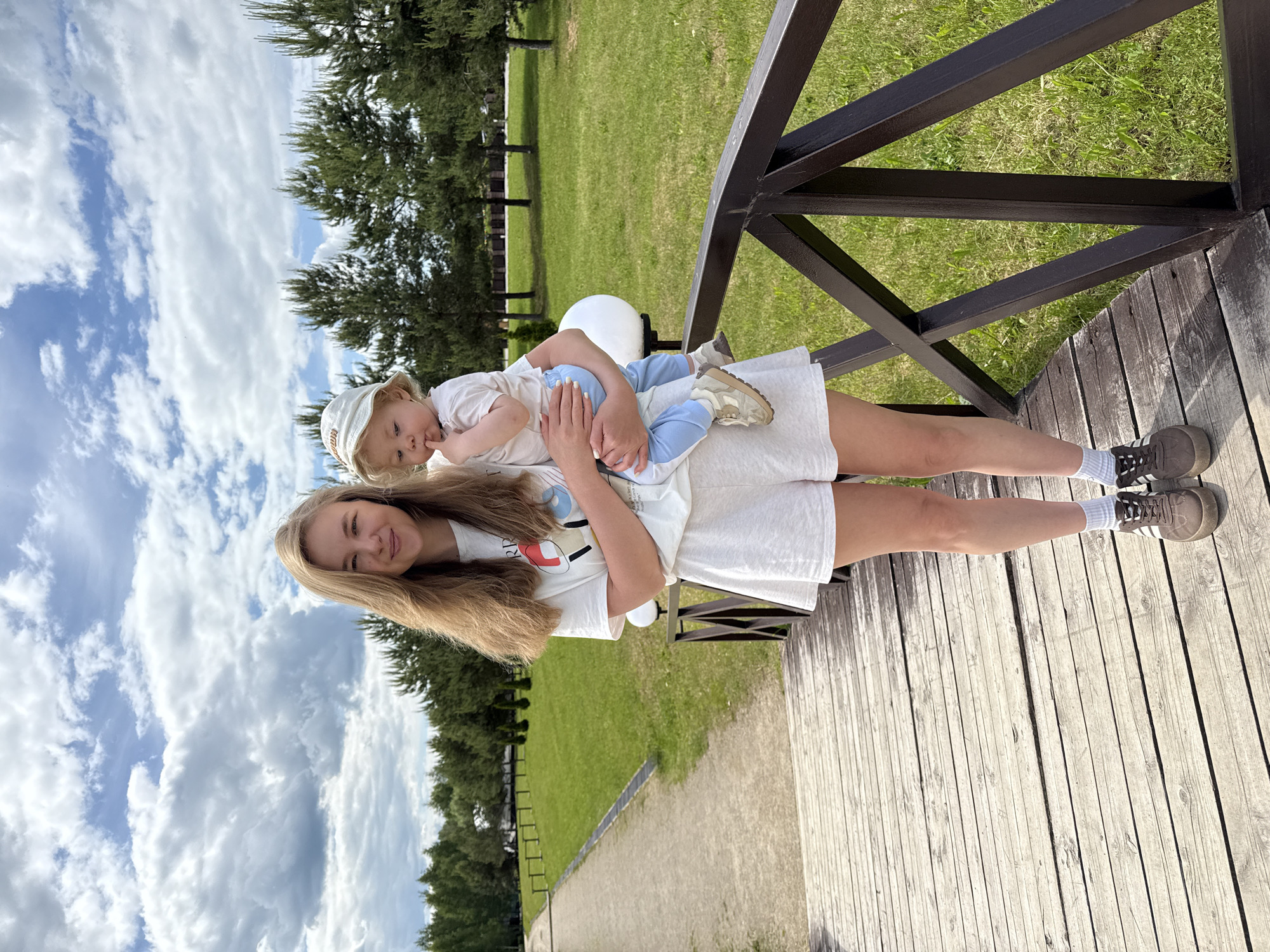 The image, correctly oriented, captures a young woman holding a baby or toddler outdoors on a bright, sunny day. The woman, with long blonde hair cascading over her shoulders, smiles gently towards the viewer. She is dressed casually in a white t-shirt with a colorful graphic print (partially showing "RP" and other abstract shapes), white shorts or a skirt, white socks, and dark grey Adidas sneakers with white stripes.
In her arms, she cradles a young child, also with blonde hair, wearing a light-colored bucket hat (with partially visible text, possibly "Mama" or "Mini"), a white top, and light blue pants, paired with small light-colored sneakers. The child appears calm, looking slightly away from the camera, possibly with a finger in its mouth.
They are standing on a light-colored wooden deck or boardwalk. To the right, a dark brown metal railing forms a diagonal element, providing a sense of depth and overlooking a vibrant green, well-maintained lawn. Beyond the lawn, a line of tall, dark green coniferous trees stands against a clear blue sky dotted with fluffy white clouds. A dirt or gravel path is visible in the distance, parallel to the treeline. The overall scene suggests a pleasant outdoor leisure moment, likely in a park or recreational area in Dobrograd, Russia, during daytime under fair weather. A small white spherical light fixture is partially visible in the mid-ground.