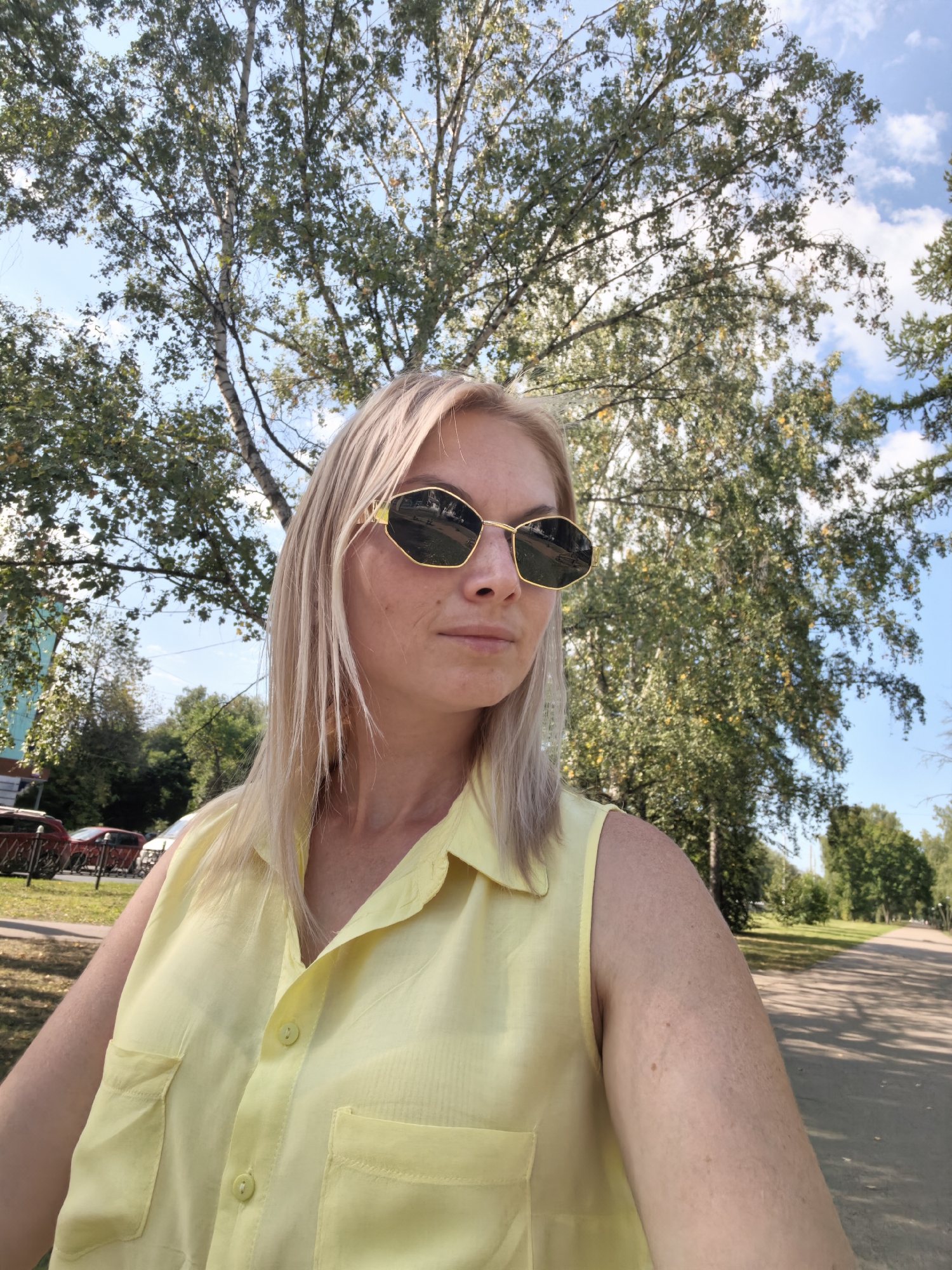 The image shows a woman with blonde hair and sunglasses, wearing a yellow sleeveless shirt, standing outdoors with trees and a blue sky in the background. The woman is positioned in the foreground, slightly off-center to the left, with her face turned to the right. Her sunglasses are reflective, showing a distorted image of the surroundings. The shirt has two chest pockets and two buttons down the front. Behind her, a tree with green foliage dominates the upper portion of the frame, with a portion of another tree to the right. The background also shows a paved path and some greenery, suggesting a park or a residential area with trees. The lighting indicates it is daytime, likely sunny. The specific location in the city of Sterlitamak, Russia, cannot be determined from the visual information provided in the image alone.