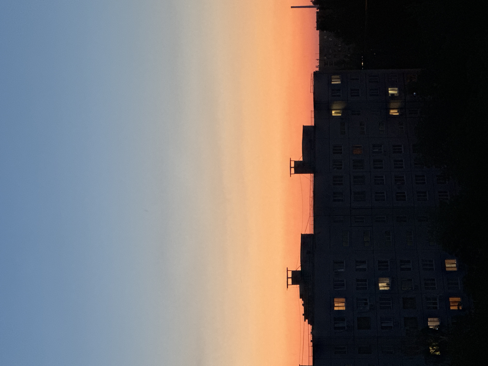 An outdoor, full-frame, portrait orientation photograph captures a dusky sky over a silhouetted apartment building. The sky transitions from a pale blue at the top to a vibrant orange and then a deep red towards the horizon, indicating either sunrise or sunset. A multi-story apartment building dominates the lower half of the frame. Its facade is dark and appears to be made of concrete or similar material. Several windows are visible, some of which are lit from within, suggesting occupancy. The top of the building features several architectural elements, including what appear to be ventilation shafts or chimneys. Dark silhouettes of trees frame the bottom of the building, obscuring the ground level. In the distance, other buildings and structures are faintly visible against the horizon, also silhouetted by the fading light. The overall scene evokes a sense of urban twilight in an unspecified location.