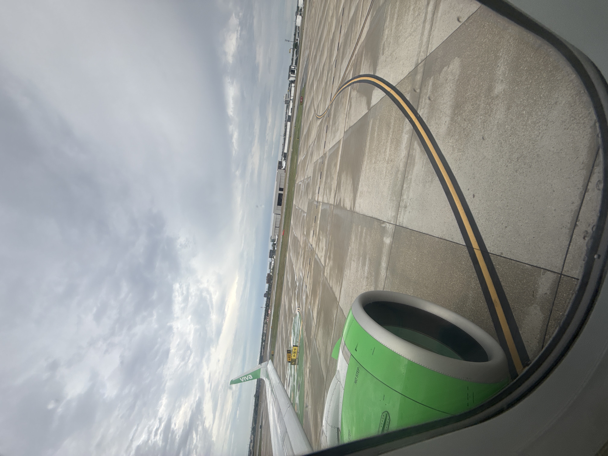 The image shows a view from an airplane window. An engine is visible in the foreground, colored green and grey. The airplane appears to be taxiing on a grey concrete surface with black and yellow painted lines. Beyond the concrete, there are buildings and other infrastructure, suggesting an airport environment. The sky is overcast with grey clouds. The plane is likely on the ground at San Antonio International Airport in the United States of America, due to the presence of Viva livery and the infrastructure of the airport.