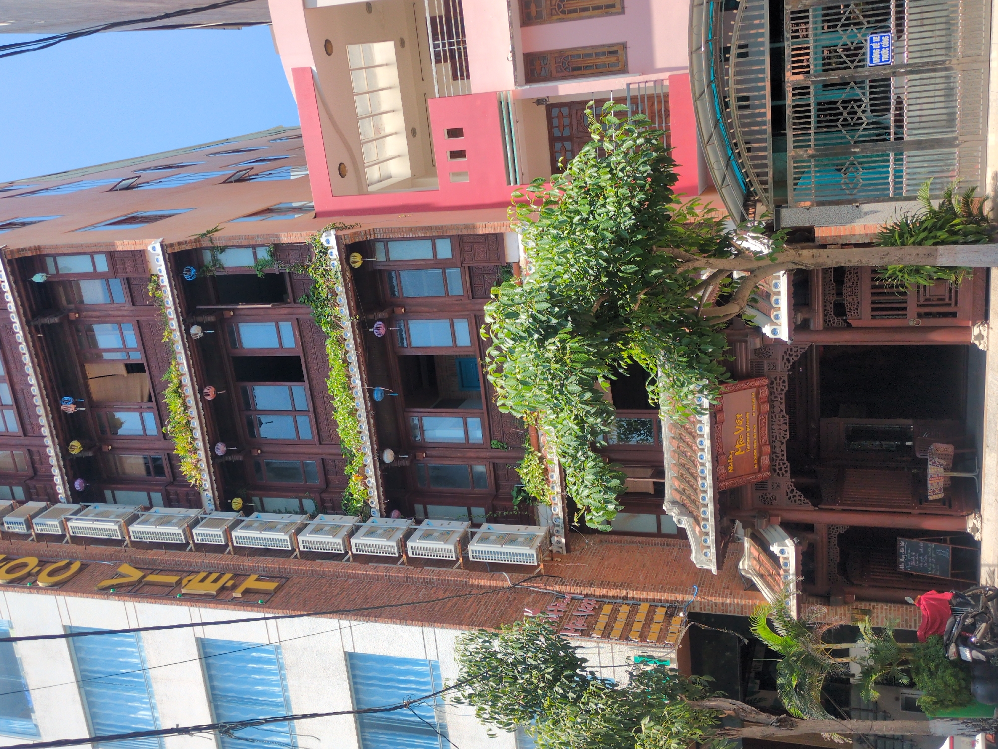 An eye-level street view depicts a dense urban environment in South Quy Nhon Ward, Vietnam, under clear daylight. The central subject is a multi-story building featuring traditional Vietnamese architecture, characterized by a dark brown wooden facade with intricate carvings, traditional red-tiled roof accents, and multiple balconies adorned with lush greenery and colorful decorative lanterns. A prominent sign on the ground floor identifies it as "Nhà hàng Mộc Việt" (Moc Viet Restaurant) and "Vietnamese Gastronomy," with a contact number. The restaurant entrance is open, revealing a dark interior, and a black chalkboard sign with Vietnamese text, along with a small table and two chairs, are positioned outside.
To the left, a tall, modern white building with numerous blue-tinted rectangular windows rises. A narrow brick column on its right edge displays a vertical yellow "MOC VIET" sign and a stack of at least eleven white outdoor air conditioning units. Overhead electrical wires are visible.
To the right of the central traditional building is a multi-story structure predominantly painted in shades of pink, featuring balconies and a large, ornate metal gate. A blue rectangular sign on the gate reads "KHÔNG ĐỂ XE TRƯỚC CỔNG" (No parking in front of gate).
Vegetation includes several trees with dense green foliage partially obscuring the buildings and potted plants near the traditional building's entrance. A single black motorbike, partially covered by a red cloth, is parked on the sidewalk in front of the traditional building. No individuals are visible in the scene.