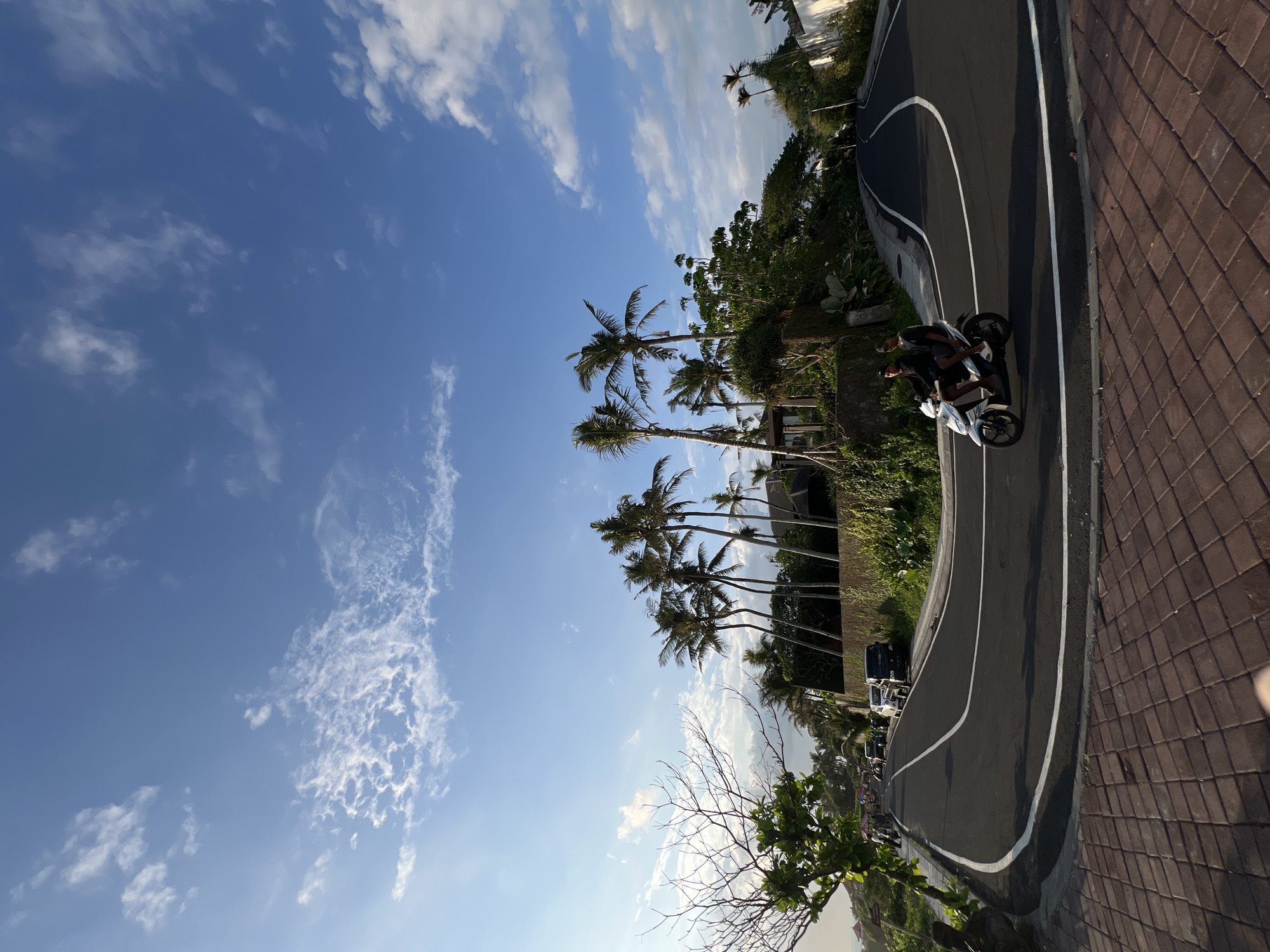 The image depicts a scene in Mengwi, Indonesia, showing a winding road with palm trees on one side and a brick pathway on the other. A motorcycle with two riders is traveling on the road. The sky is blue with scattered clouds. Other vehicles can be seen parked along the road.