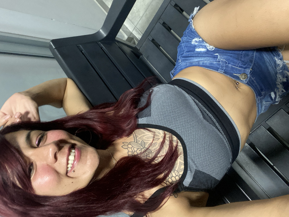 A woman with dark red hair is lying on a black bench. She is wearing a gray sports bra with black trim and ripped denim shorts. Her navel is pierced, and she has a tattoo on her chest. She is smiling, revealing a pierced lip. The bench appears to be outdoors, possibly on a patio or deck.