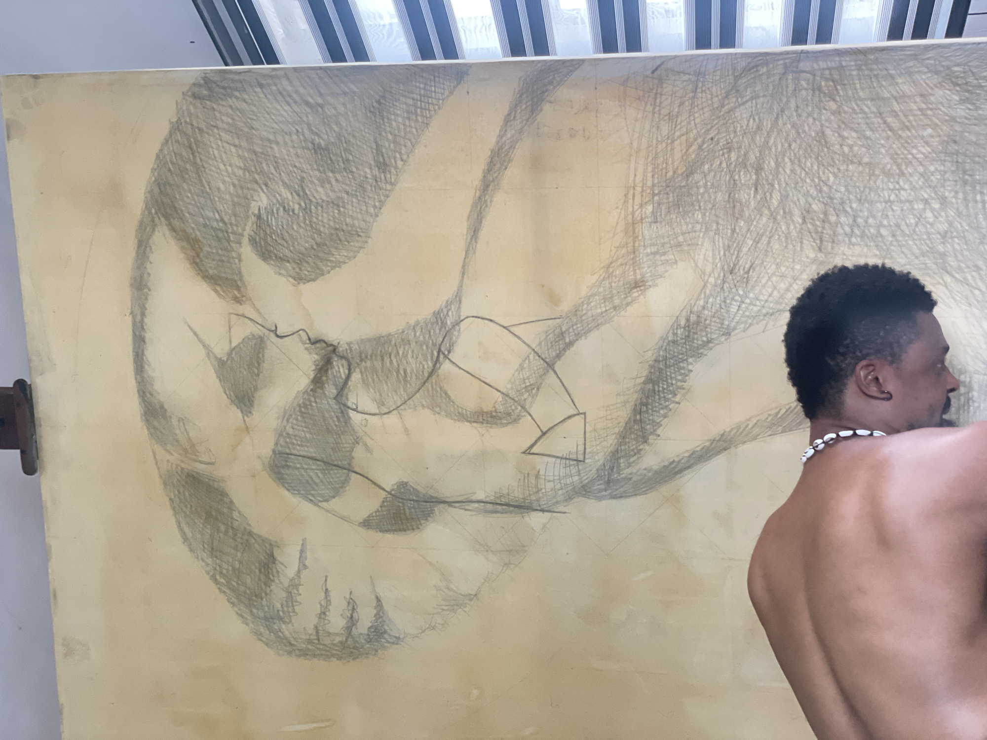 The image displays an indoor scene, likely an art studio or gallery in Ado, Nigeria, featuring a man beside a large artwork. An adult male, seen from behind and in profile on the right side, is the human subject. He has dark skin, short dark curly hair, and a well-defined, muscular back. He wears a distinctive necklace made of white shells and a small dark earring in his visible ear. His head is turned to the left, suggesting he is looking towards the artwork or past it.
The central subject is a large, light yellowish-brown drawing, possibly on paper or canvas, mounted on an easel or propped up. The artwork is executed in pencil, featuring extensive areas of dark gray hatching that create textured, volumetric forms. These forms appear abstract or highly stylized, possibly suggesting organic shapes or a skull-like structure, with some elements resembling small, pointed trees towards the bottom left. Interspersed within the broad shaded areas are sharper, more geometric lines, including arrow-like shapes, indicating a dynamic or directional quality. Faint grid lines are subtly visible beneath the drawing, suggesting a preparatory stage or a method for scaling the artwork.
The background at the top of the image reveals a window with horizontal blinds or slats, allowing bright light into the space, indicating daytime. The scene conveys a sense of artistic creation or contemplation. No clear, legible text is visible within the image, though faint, indistinct markings can be seen on the artwork's surface.