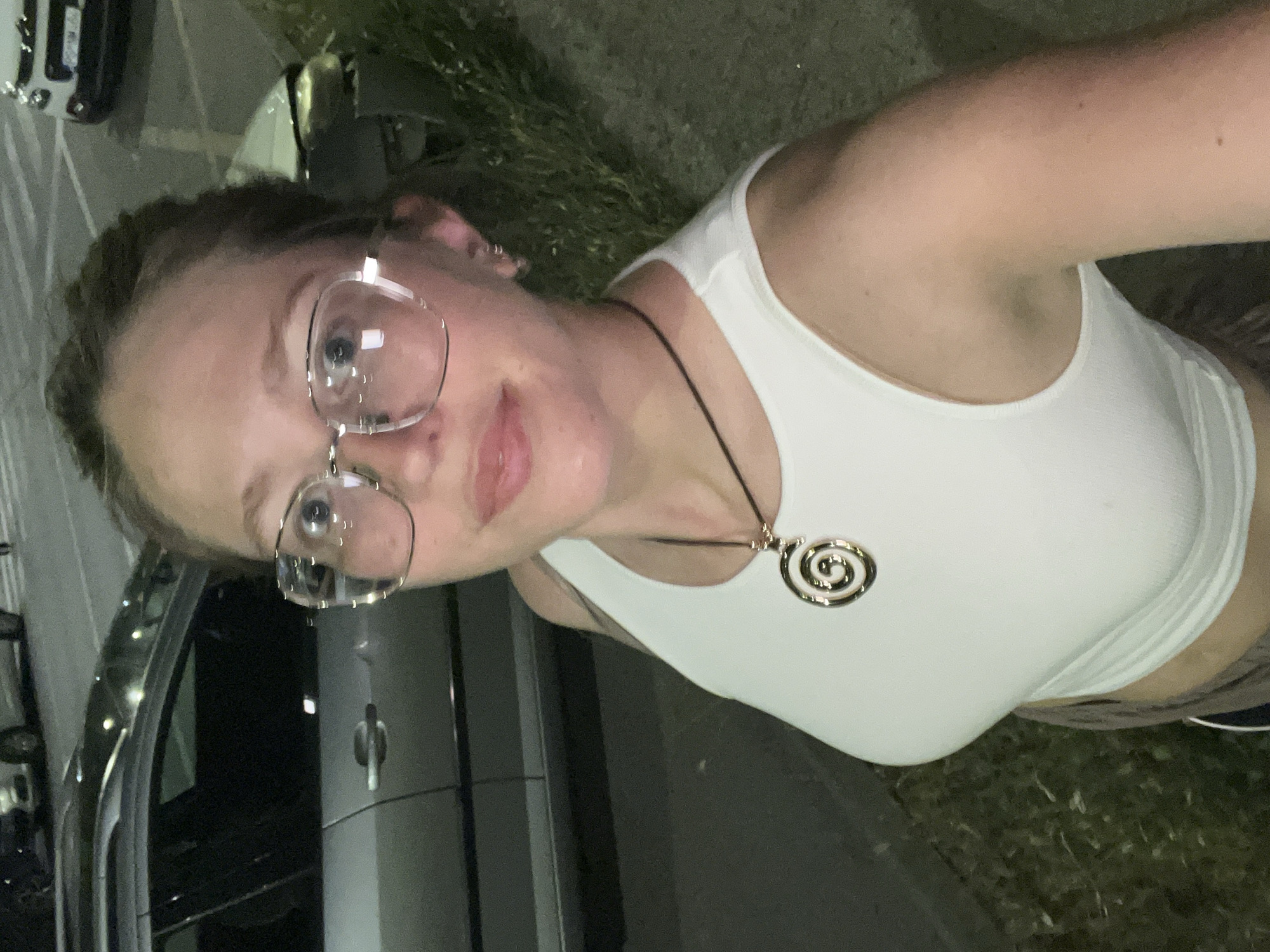 A woman with light skin and hair pulled back, wearing clear-framed glasses, a white tank top, and a necklace with a circular pendant, stands outside at night. She is positioned near a gray car, with other vehicles visible in the background. Grass is visible near the vehicles, and the lighting suggests it is in an urban or suburban setting, likely Podgorica, Montenegro, though no specific landmarks confirm the location.