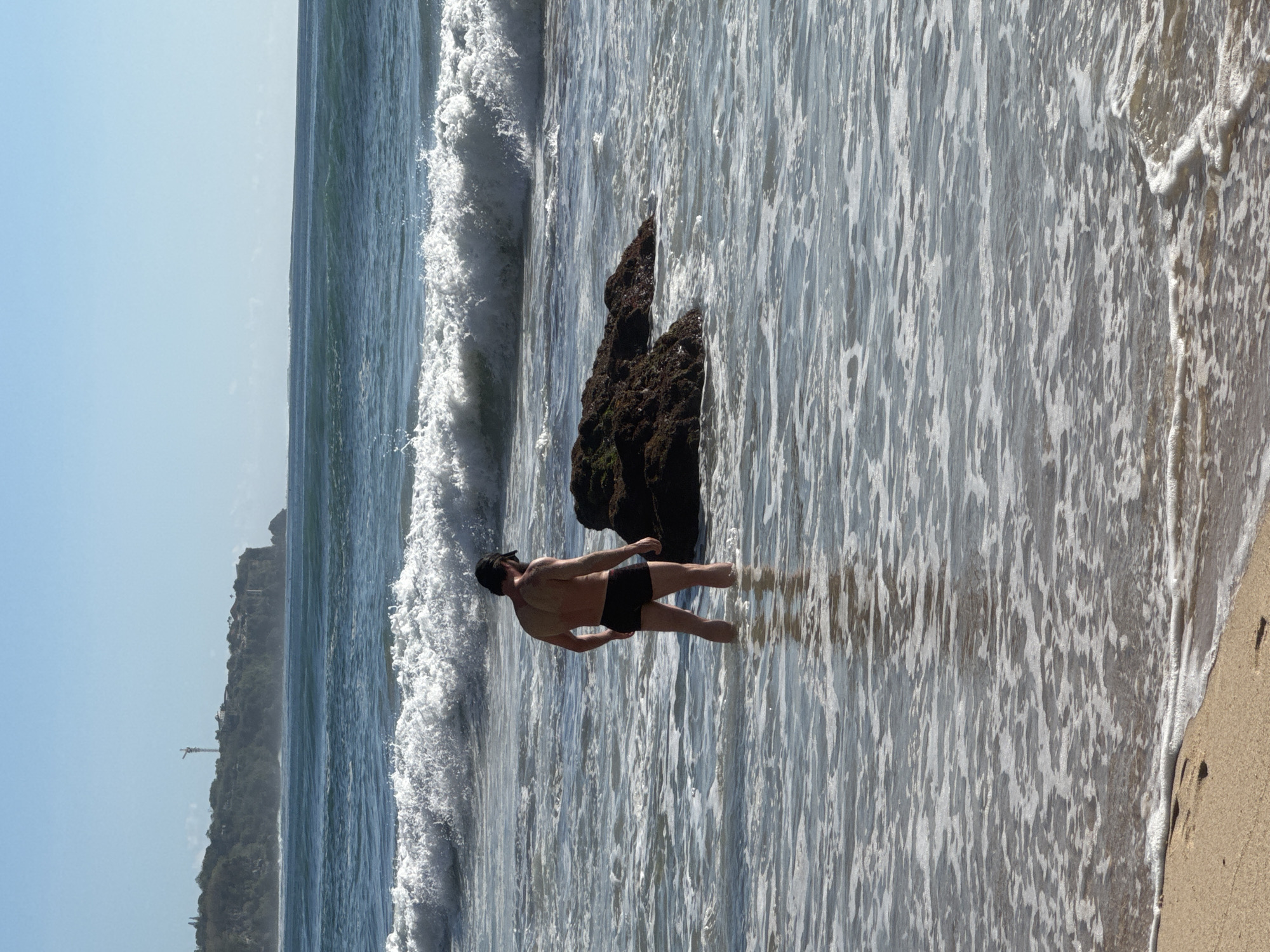 A man wearing black swim trunks and a black baseball cap stands in shallow ocean water on a beach. He is facing away from the camera, looking towards a large rock formation protruding from the water. Behind the man, waves are crashing on the shore. In the distance, a cliffside with vegetation and buildings is visible, topped by a tall tower with a crane. The sky is clear and blue. The scene depicts a coastal environment in Kuta Selatan, Indonesia.