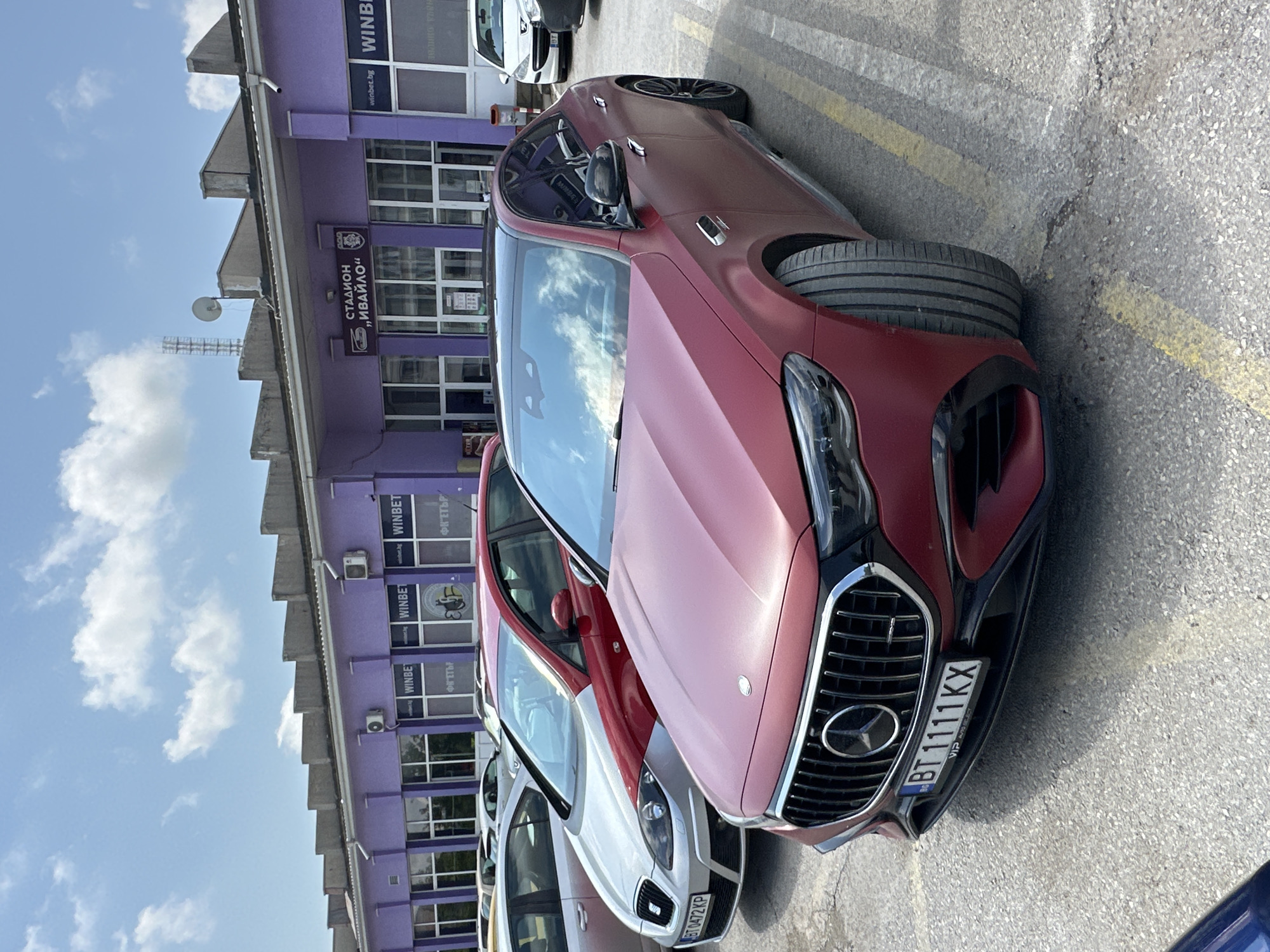 The image, rotated 90 degrees clockwise, captures an outdoor scene under a bright, partly cloudy sky. Several cars are parked on an asphalt surface marked with yellow lines, facing a prominent, long, purple building.
In the immediate foreground, a matte dark red Mercedes-Benz sedan, likely an AMG model, is visible. Its Bulgarian license plate reads "BT 1111 KX". Behind it, a white car, possibly a SEAT, is partially visible, bearing the license plate "BT 0472 KP". Other vehicles are parked further down the row.
The purple building in the background features multiple windows and various signs. Several signs display "WINBET" and "winbet.bg", indicating a betting company. Another sign reads "ФК ЕТЪР" (FC Etar), suggesting a football club. A large sign prominently displays "СТАДИОН „ИВАЙЛО"" (Stadium "Ivaylo"), confirming the location as Ivaylo Stadium in Veliko Tarnovo, Bulgaria. Small air conditioning units and a satellite dish are visible on the building's exterior, and a tall antenna structure rises above the roofline in the distance.
The scene depicts a typical daytime setting, likely mid-day, with clear weather. There is no specific activity or event taking place other than parked cars at a stadium complex.