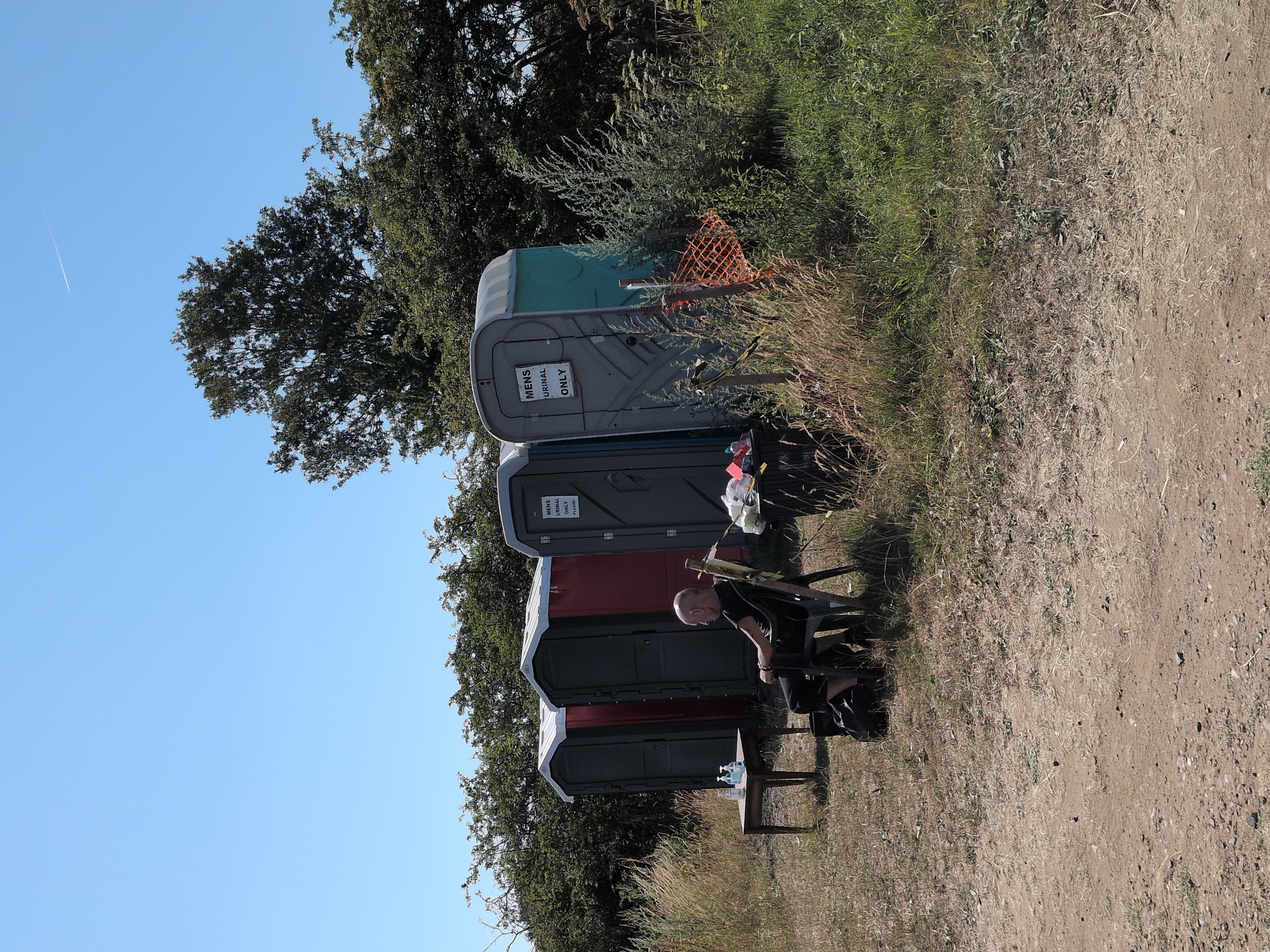 A man sits in a black chair outside several portable toilets under a clear blue sky in Claverley, United Kingdom. He is bald and wearing a black t-shirt and pants. In front of him is a small wooden table with two bottles of hand sanitizer. To his right, there is a large black trash can with some items in it. Behind him, there are four portable toilets. The first is grey, the second is red, the third is grey and labeled "MENS URINAL ONLY PLEASE", and the fourth is teal and grey. There is tall, dry grass and green bushes surrounding the area, with trees in the background. The ground is dirt and gravel.