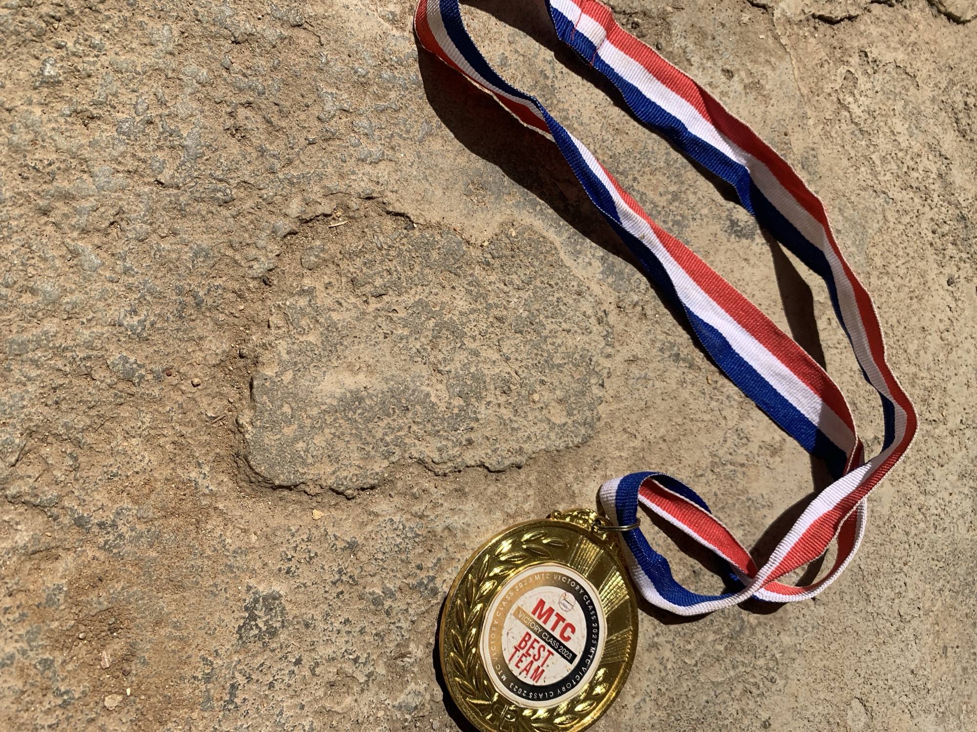 A gold medal with a red, white, and blue striped ribbon is depicted lying on a rough, textured ground surface in Rafin Par, Nigeria. The circular gold medal features an embossed laurel wreath design encircling its perimeter. The outer ring of the medal bears the inscription "MTC VICTORY CLASS 2023" repeated in a circular pattern. The central design includes a smaller white circle with "MTC" in prominent red text, positioned directly above "VICTORY CLASS 2023" and "BEST TEAM" in black and red lettering.
The ribbon, woven with distinct red, white, and blue vertical stripes, is looped and partially extends out of the frame to the left. It connects to the medal via a small gold ring. The ground surface is composed of a weathered, concrete-like material, characterized by an uneven texture, visible cracks, and scattered fine soil and small pebbles. The color of the surface varies, displaying shades of tan, brown, and gray. Strong, direct sunlight illuminates the scene, casting sharp shadows from the ribbon and the right side of the medal onto the ground, indicating an outdoor environment. No individuals, actions, or direct interactions are visible within the frame.