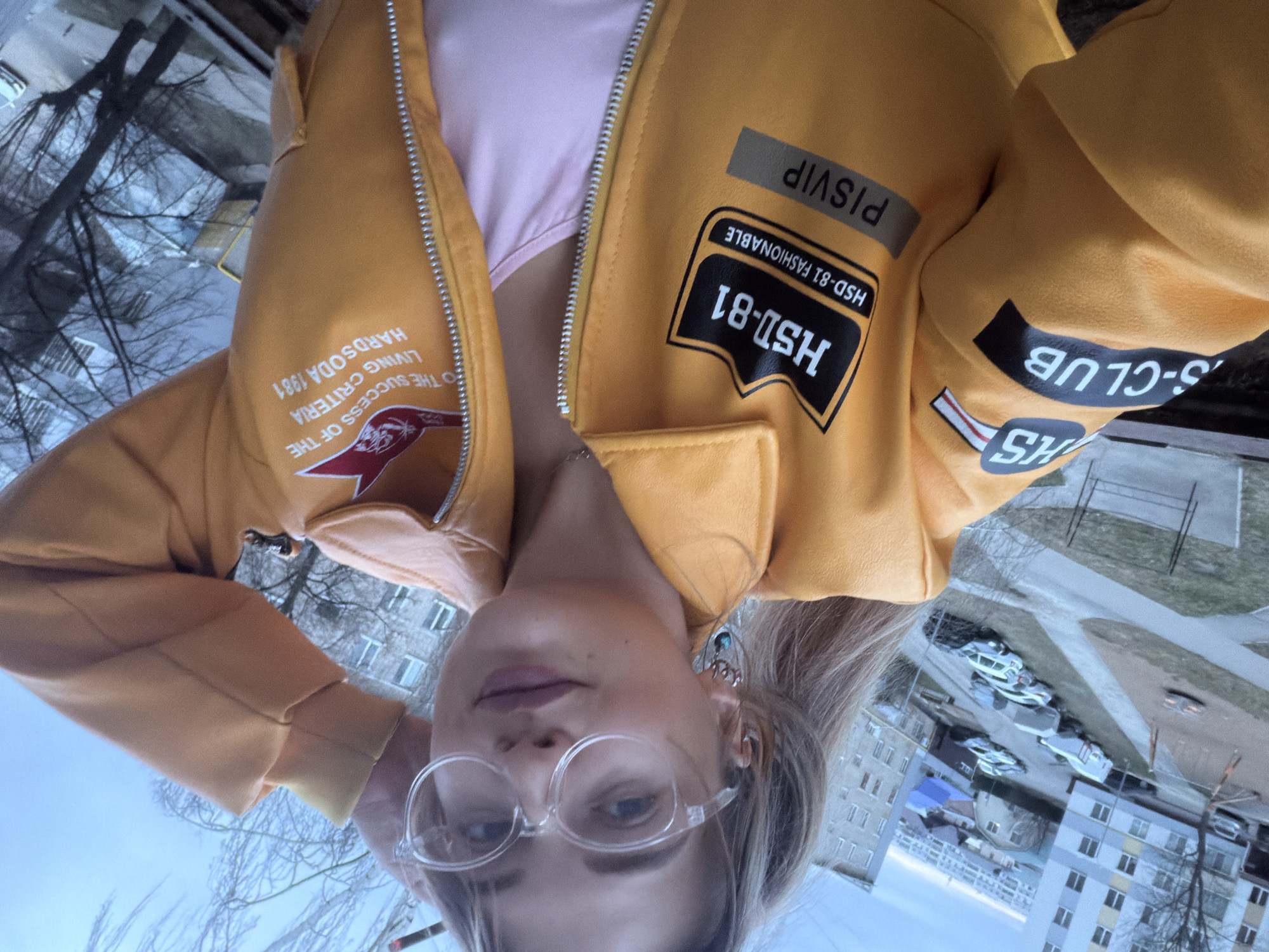 The image shows a person wearing glasses and a yellow jacket with patches and lettering, including "HS", "S-CLUB", "HSD-81", "HSD-81 FASHIONABLE", "PISVIP", and "THE SUCCESS OF THE LIVING CRITERIA HARDSODA 1981". The jacket is partially open, revealing a light pink garment underneath. The background shows a city scene with trees, buildings, and cars. The location is likely Naberezhnye Chelny, Russia, based on the urban environment typical of Russian cities.