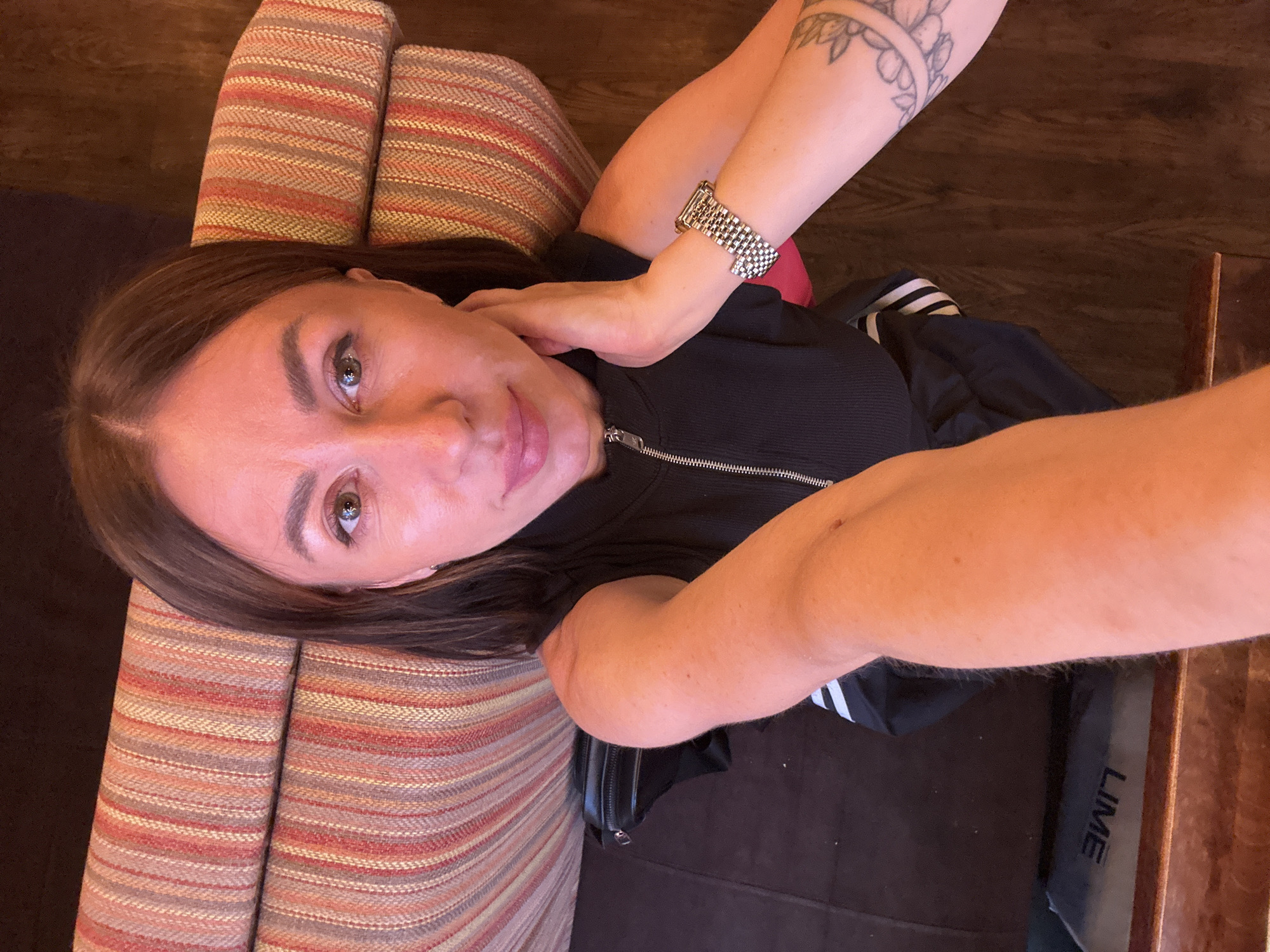 A female subject, with medium-brown hair and light makeup, is depicted in a high-angle selfie. She wears a black, short-sleeved top with a silver front zipper. A metallic wristwatch with a linked band adorns her left wrist, while a floral or decorative tattoo is visible on her right forearm. Her left hand is positioned against her neck/chin. She is seated on upholstery featuring vertical stripes in shades of red, orange, beige, and grey. To her left, a black fanny pack rests against the cushion. The flooring to her right is dark wood. An item on the floor, near her lower left, displays the word "LIME" in white capital letters, oriented upside down. The background consists of dark, indistinct surfaces. This scene is located in the city of Kazan, Russia.