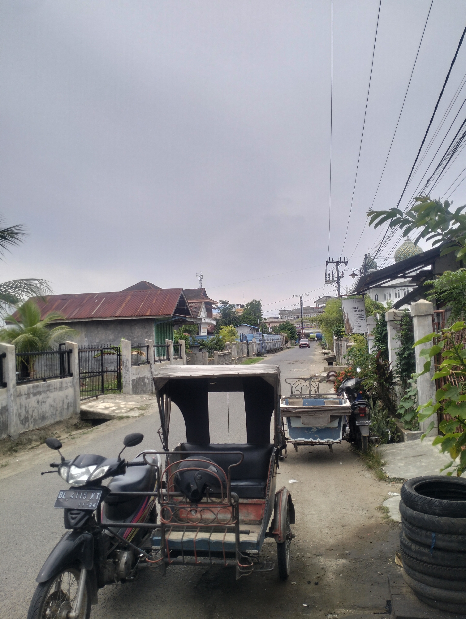 A cloudy day in Lhokseumawe, Indonesia, shows a residential street scene. In the foreground, a black motorcycle with a sidecar is parked on the asphalt road. The motorcycle's license plate reads "BL 4115 KT". The sidecar is red and black with a black helmet inside. Further down the road, another motorcycle with a sidecar is parked. Residential houses with corrugated metal roofs line both sides of the street. One house has a gray concrete wall with a black metal fence. A mosque with a green dome is visible in the distance. Power lines crisscross the sky. Lush green foliage is present on the right side of the image.