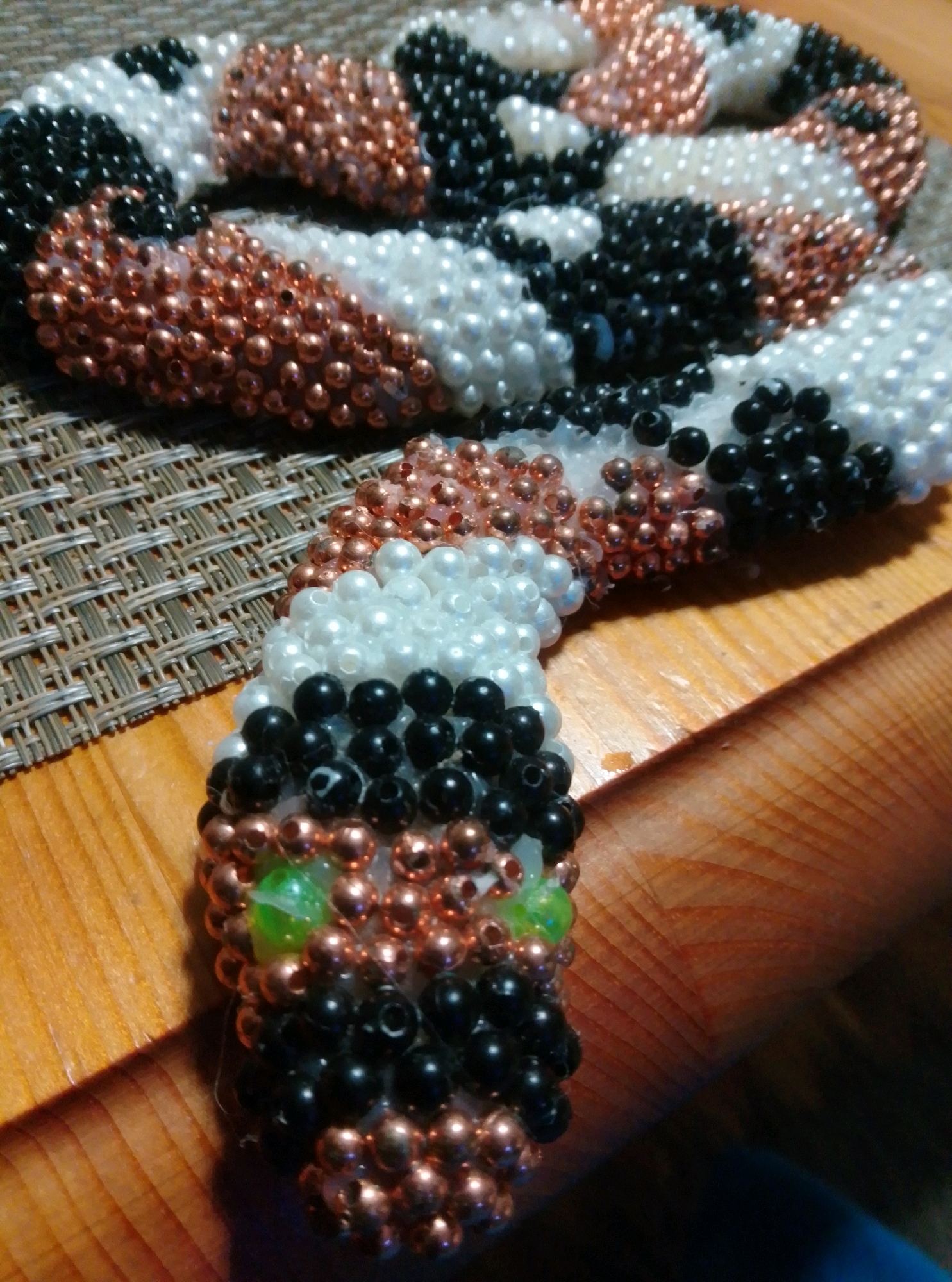 A beaded snake lies on a woven placemat on a wooden surface. The snake is coiled and appears to be made of white, black, and copper-colored beads. The head of the snake is visible in the foreground, with two green beads for eyes. The background is out of focus. This image was taken in the city of Wedel, Germany.