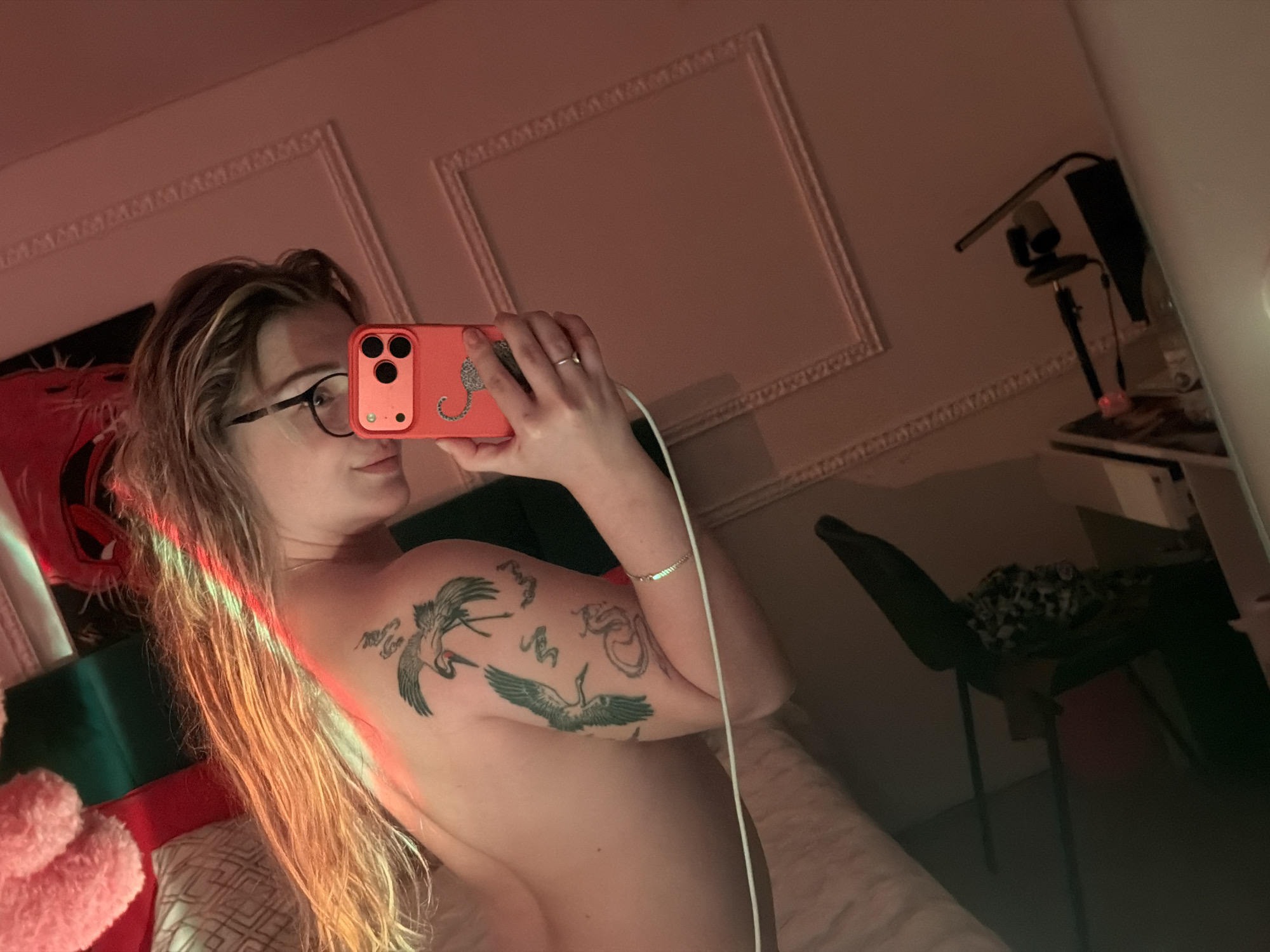 A medium mirror selfie captures a young woman with long, light-colored hair and glasses, holding a coral/orange smartphone with a distinctive leopard-print case. She appears partially nude, with her bare left shoulder and arm prominently displaying tattoos of two cranes and other abstract designs. Her right hand, adorned with a ring and a thin bracelet, holds the phone, which has a white charging cable attached.
The scene is an indoor domestic setting, likely a bedroom, viewed through the mirror's reflection. The room is dimly lit with a dominant red/pink ambient light, casting a warm, moody glow on her hair and skin. Visible in the reflection are decorative white molding on the wall, a black chair, and a standing light fixture or camera setup. In the lower left, a red object, possibly a cushion or stuffed animal, and a pillow with a geometric pattern are visible. The lighting suggests it is evening or night. The woman's expression is neutral and relaxed as she takes the photo. No discernible text is visible on any objects, though the tattoos feature stylized elements. This scene is contextualized as being in Большая Шильна, Russia.
