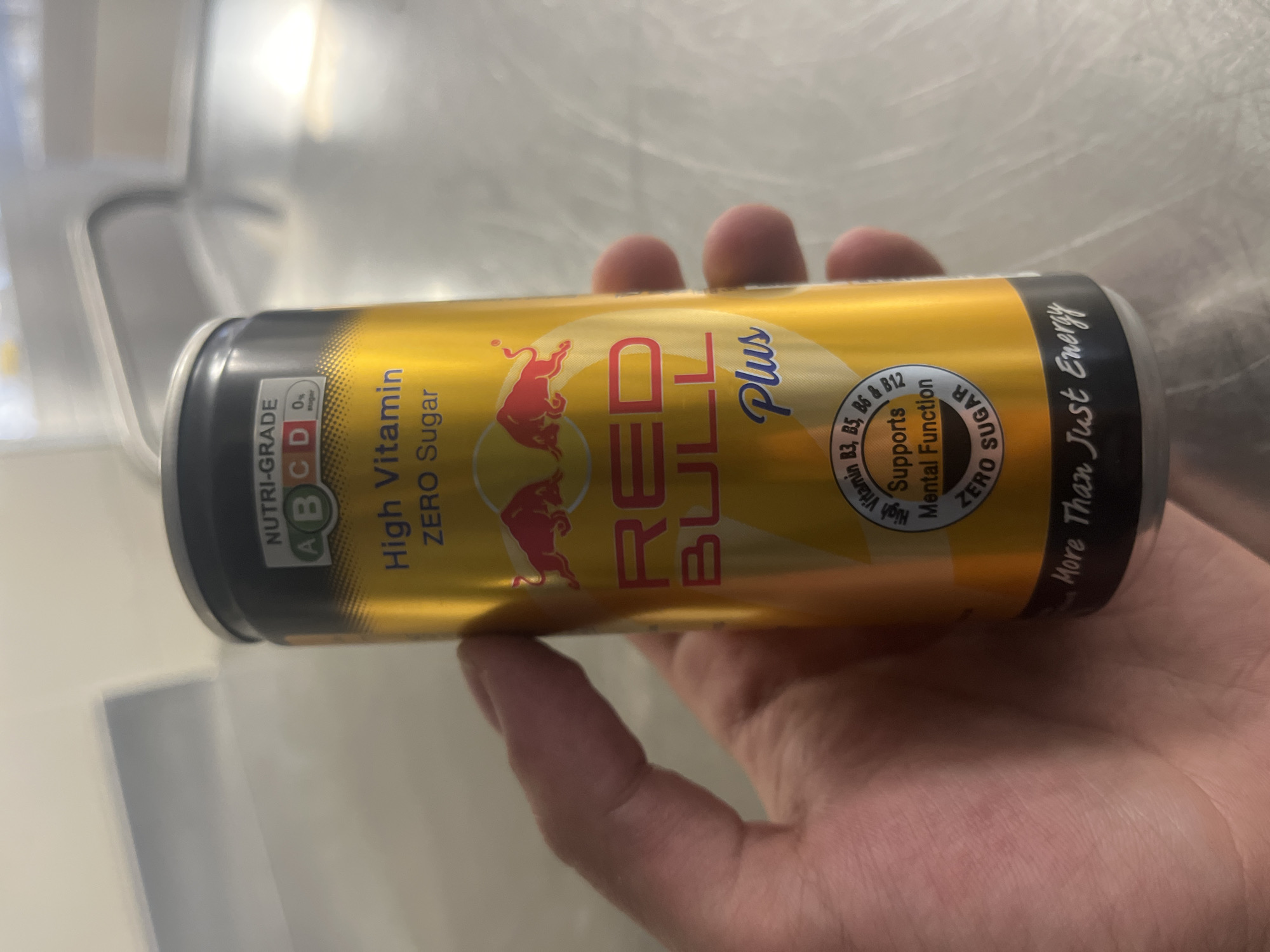 A hand is holding a can of Red Bull Plus Zero Sugar energy drink. The can is gold colored with a silver top and bottom. The text "High Vitamin ZERO Sugar" is printed on the can. There is a logo of two red bulls facing each other. The text "RED BULL Plus" is in red. The can also says "Supports Mental Function Vitamin B3, B5, B6 & B12", "ZERO SUGAR" and "More Than Just Energy". The NUTRI-GRADE is ABCD. The background is a blurred silver surface. It is not possible to ascertain the location of this image as Sofia, Bulgaria.