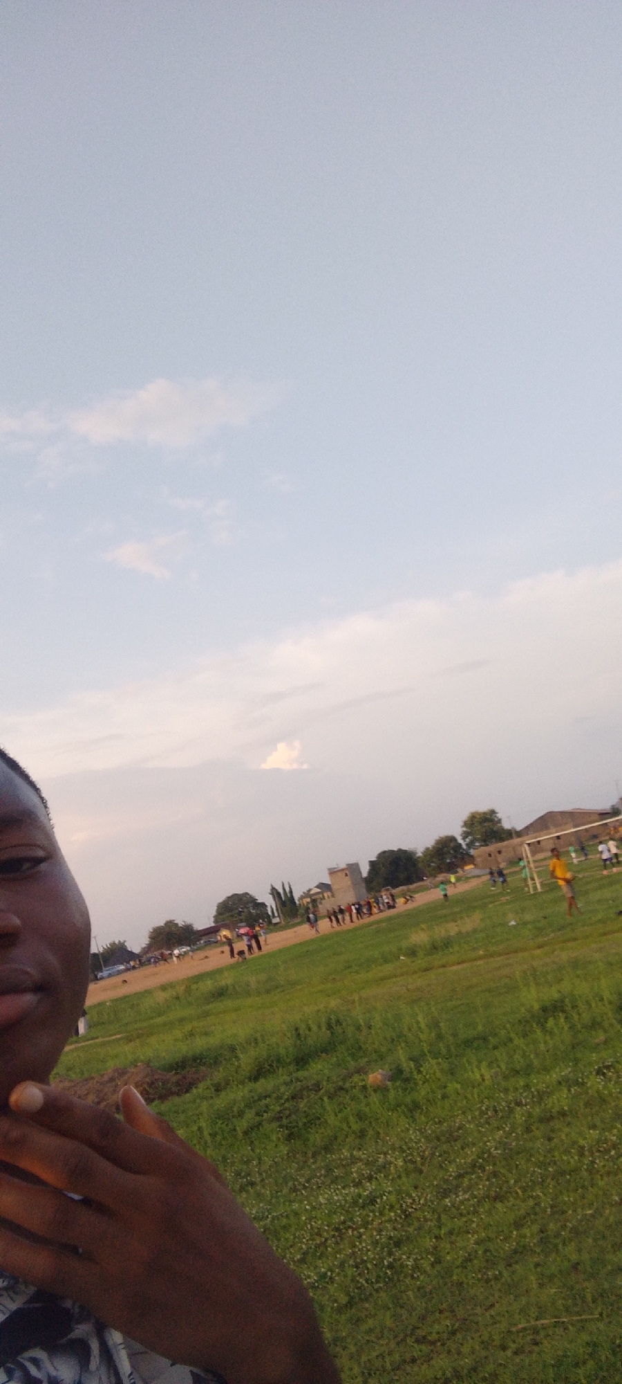 The image presents a selfie of a dark-skinned person, with part of their face and hand visible in the lower left corner. The background shows a grassy field with people gathered, some appearing to play soccer, indicated by the presence of a goal post. A building is visible further back, in the background. The sky is partially cloudy. The location appears to be in Ibo, Nigeria, inferred from the query context.