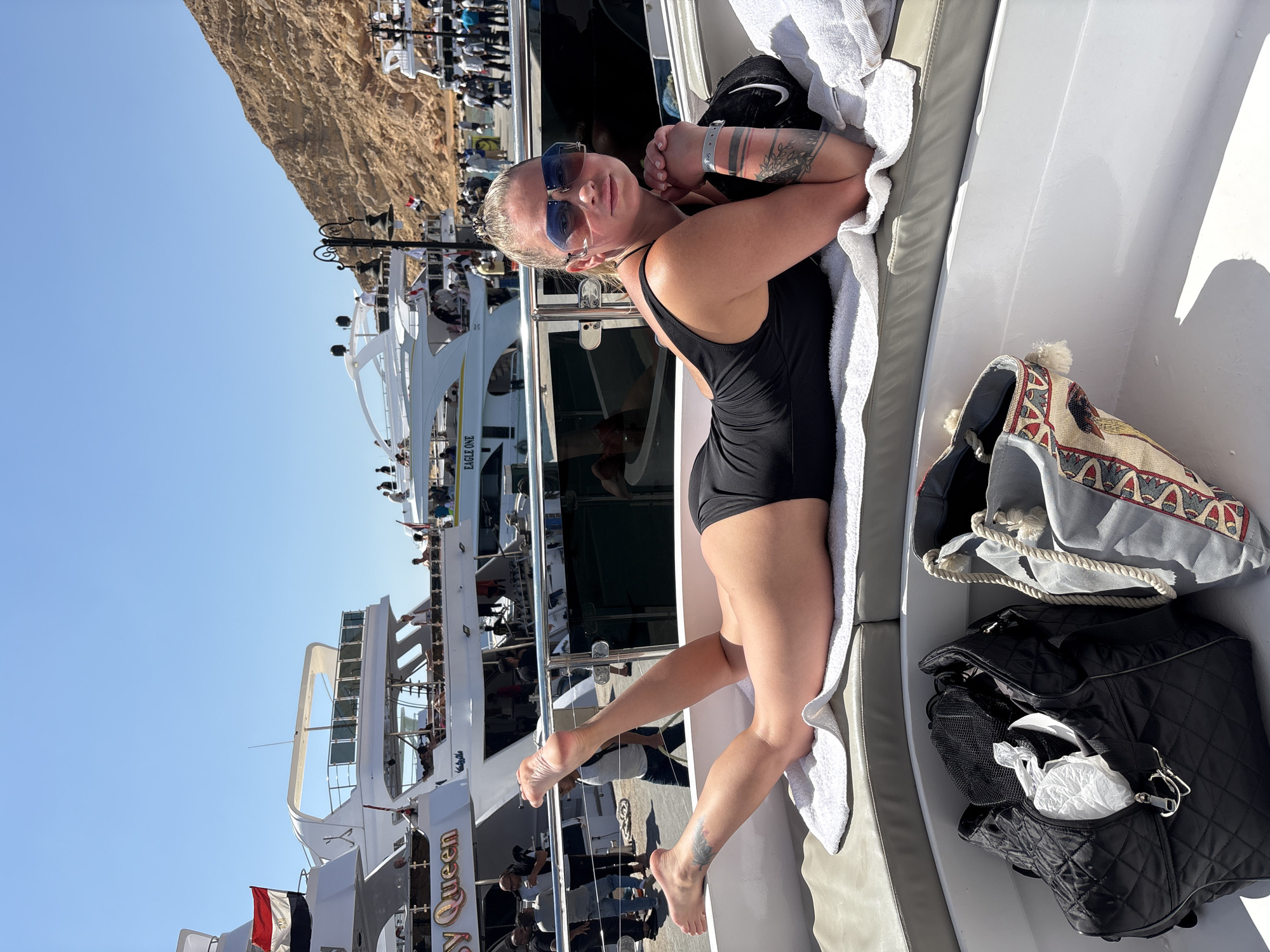 A woman wearing a black swimsuit and blue sunglasses is lying on a white lounge cushion on a boat or yacht. She has tattoos on her arms and foot. Beside her are a white towel and a couple of bags. In the background, other boats are docked, with people visible on them. A rocky hill rises up in the background. The boat she is on appears to be docked in Sharm El Sheikh, Egypt.