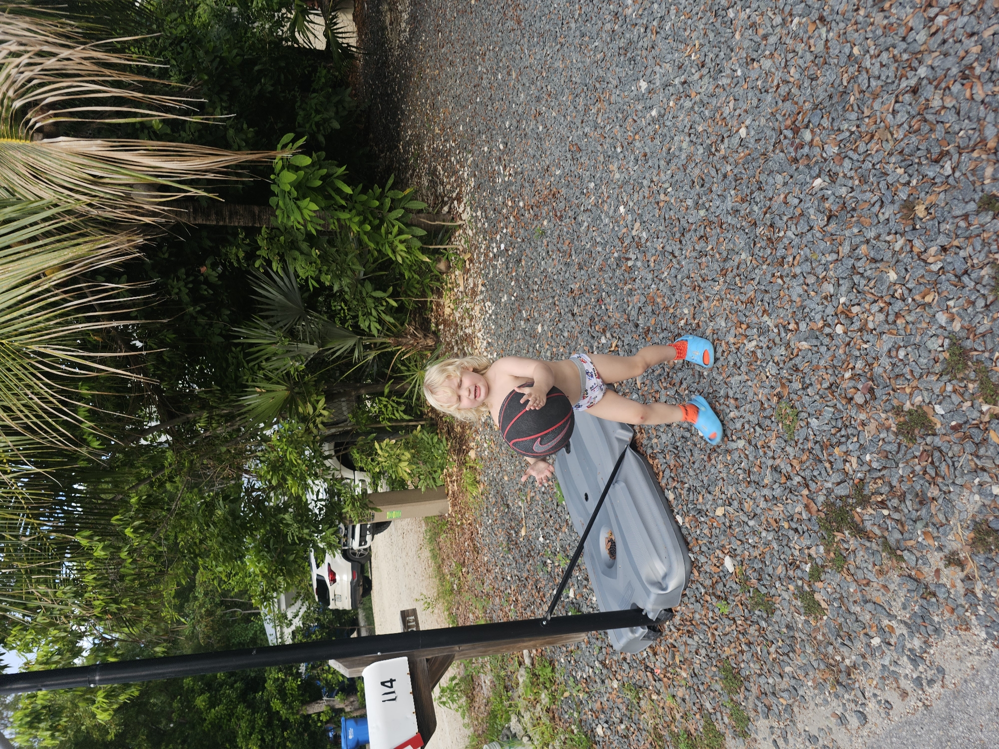 A fair-skinned child with blonde hair stands on a gravel driveway holding a basketball. The child is wearing swim trunks and blue sandals. Behind the child is a portable basketball hoop with a gray base. A mailbox with the number 114 is to the left of the child. In the background, there are trees, palm trees, and two white SUVs. The scene is outdoors, possibly in a residential area in Islamorada, Florida.