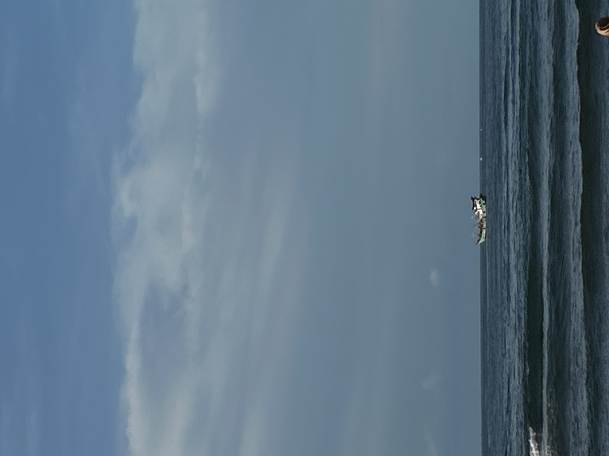 The image shows a seascape at Lhokseumawe, Indonesia. The right side is occupied by choppy sea with visible waves. A small vessel with several people can be seen in the water. The left side and the top of the image contain a light blue sky with scattered white clouds. A person's head is visible in the upper right corner.