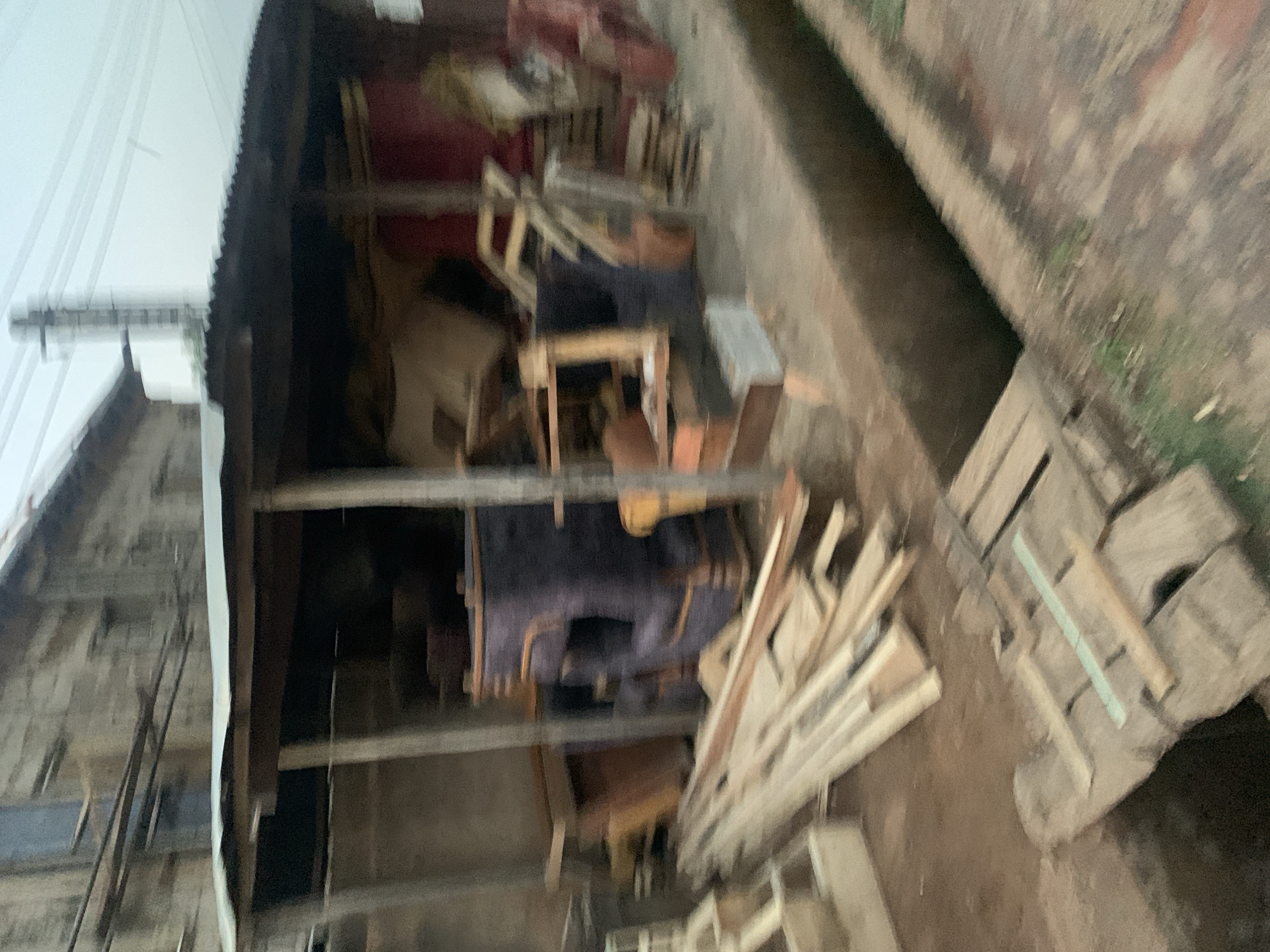 The image depicts a cluttered and disorganized outdoor storage area. It appears to be adjacent to a building, possibly a warehouse or workshop, with a partially visible corrugated metal roof. Inside the storage area, there is an assortment of furniture, including what appears to be sofas and chairs, mixed with lumber, wooden planks, and other construction materials. The ground is uneven and covered with debris. The overall impression is one of neglect and disuse.
The context of Kabong, Nigeria is not specifically evident within the image itself, but the scene suggests a location where storage space may be limited or under-resourced. There are no people visible in the frame.