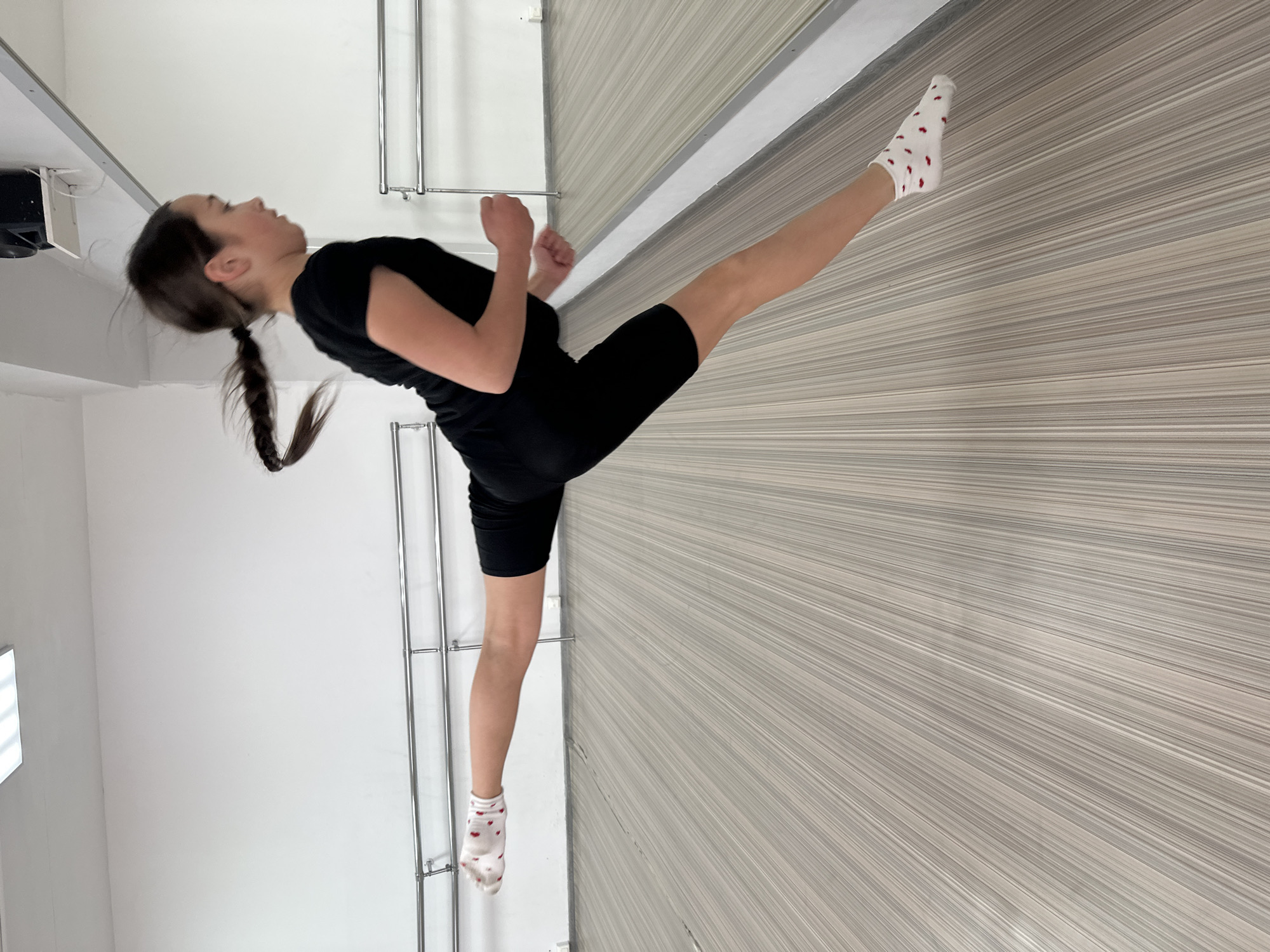 A young woman wearing a black t-shirt, black shorts, and white socks with red hearts is shown in what appears to be a dance or exercise studio. She is in a kicking pose, with one leg extended towards a mirrored wall and the other leg supporting her. Her hair is pulled back into a ponytail. The background consists of a white wall with a clothing rack and a mirror with a gray wall on the other side. The location is unknown.