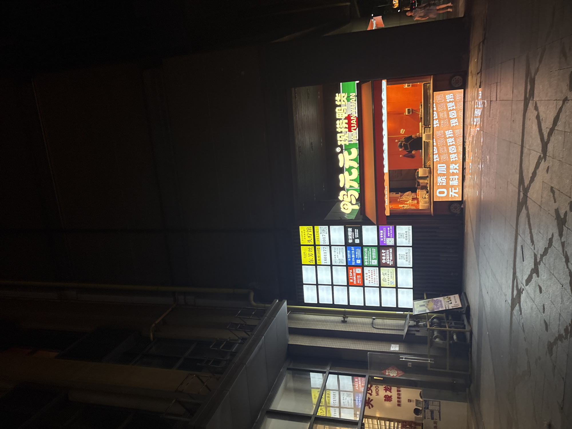 This is a nighttime photograph taken at street level, with the camera positioned looking upwards at a slight angle. The scene depicts a bustling commercial street in Haizhu District, China, illuminated by bright neon signs and storefront lights.
Dominating the center of the image is a brightly lit establishment with a large sign featuring Chinese characters and the English "YA YUAN YUAN," suggesting a business name. The sign is predominantly green and yellow. To the right of this, another sign, also in Chinese, is vertically oriented, with the text "0 添加现卤现捞" repeated multiple times, indicating a food business specializing in freshly stewed ingredients. This sign is lit with a bright white light. Further to the right, a display of smaller illuminated signs advertises services such as "SPA" and "头疗馆" (head spa), alongside phone numbers and other business details. The ground in front of these establishments is wet and reflects the light from the signs, suggesting recent rain or cleaning.
In the upper part of the image, faint figures of people can be seen walking on a raised walkway or balcony above the street-level shops. One sign on a building further back reads "美度酒店管理" (Meidu Hotel Management). The overall impression is one of a lively, commercial area active at night. The lighting creates a somewhat dramatic and vibrant atmosphere, typical of urban nightscapes.