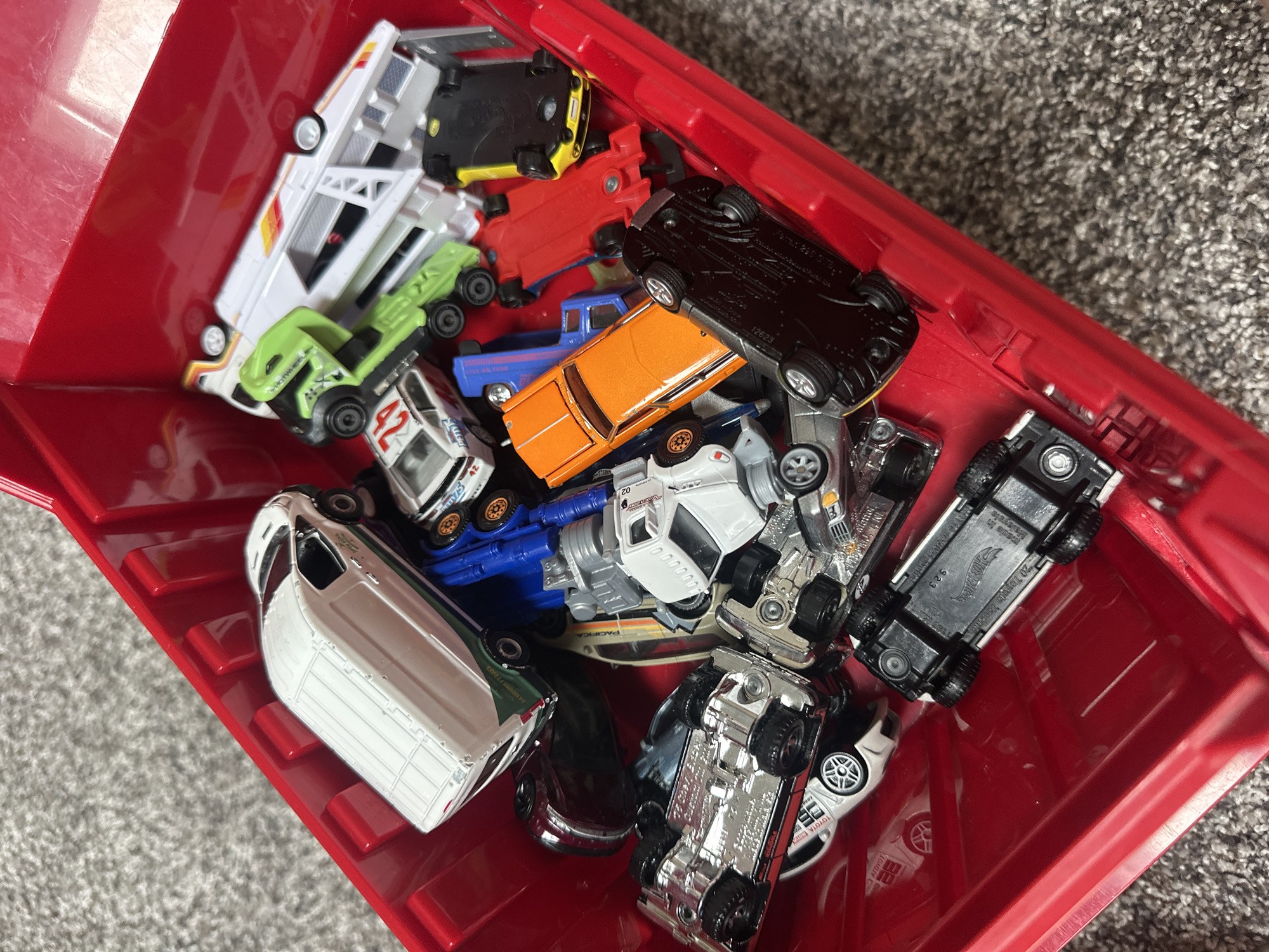 A high-angle, medium shot shows a collection of toy cars piled inside a red plastic bin. The bin is situated on a grey, textured surface that appears to be carpet. The toy cars, of various colors and types, are scattered and some are upside down. Notable vehicles include a white van with green and blue accents, a green truck, an orange car, a black car, a red car, and several silver cars. The lighting is natural and casts minimal shadows. There are no people or animals visible in the image. The scene is devoid of any context that would suggest a specific location within Schaumburg, United States of America, or any other city.