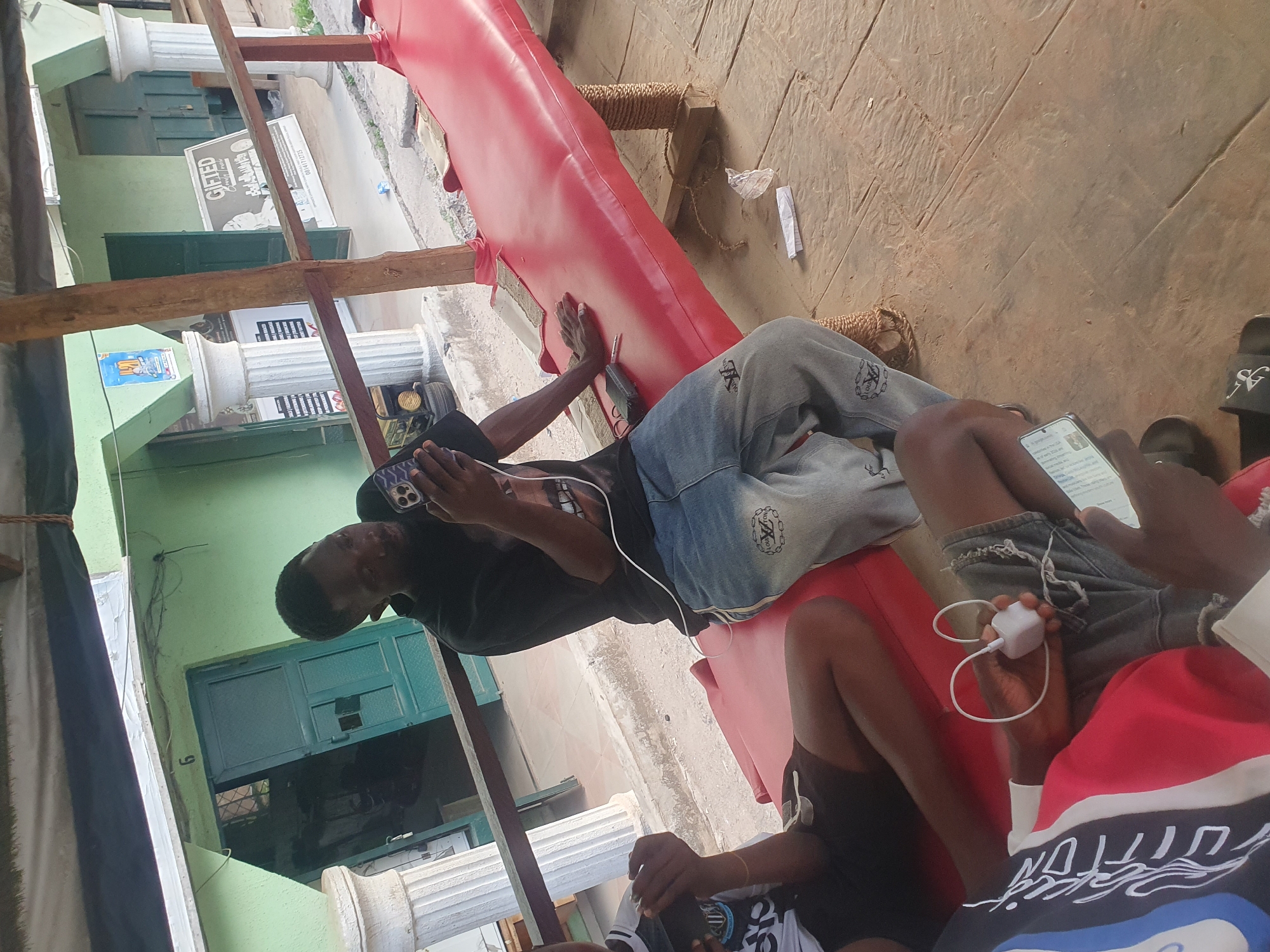 This image, captured in Lokoja, Nigeria, portrays a daytime scene of individuals relaxing outdoors, primarily engaging with their mobile phones.
In the center, a man is lying on his side on a bright red, padded bench or platform. He is wearing a black t-shirt and blue jeans, holding a smartphone with both hands as he looks at its screen. The red surface, which appears to be vinyl or leatherette, is supported by wooden posts wrapped with rope.
In the foreground on the right, two other people are partially visible. One individual is seated or reclined, holding a smartphone to which a white charging brick and cable are connected. This person wears dark shorts and a distinctive red, white, and blue hooded top, with some visible text that could be "SOLUTION." Beside them, another person's legs are seen, clad in light grey sweatpants bearing a logo similar to "CK" or Calvin Klein, and dark sandals with a white "SG" or "SIGMA" logo. This individual also appears to be focused on a mobile phone.
The setting is an outdoor or semi-covered area with a light brown, patterned concrete or tiled ground. To the left, a light green building facade is prominent, featuring white decorative pillars and dark green doors and windows, one of which has a small broken pane. Overhead, several wooden beams form part of a structure, possibly an awning. Various signs are affixed to the building: a black and white sign clearly reads "GIFTED," with additional, largely illegible text below it, and a blue and white poster is visible further along the wall. A crumpled piece of white paper and a clear plastic wrapper are discarded on the ground near the red bench. The general atmosphere is casual and relaxed.