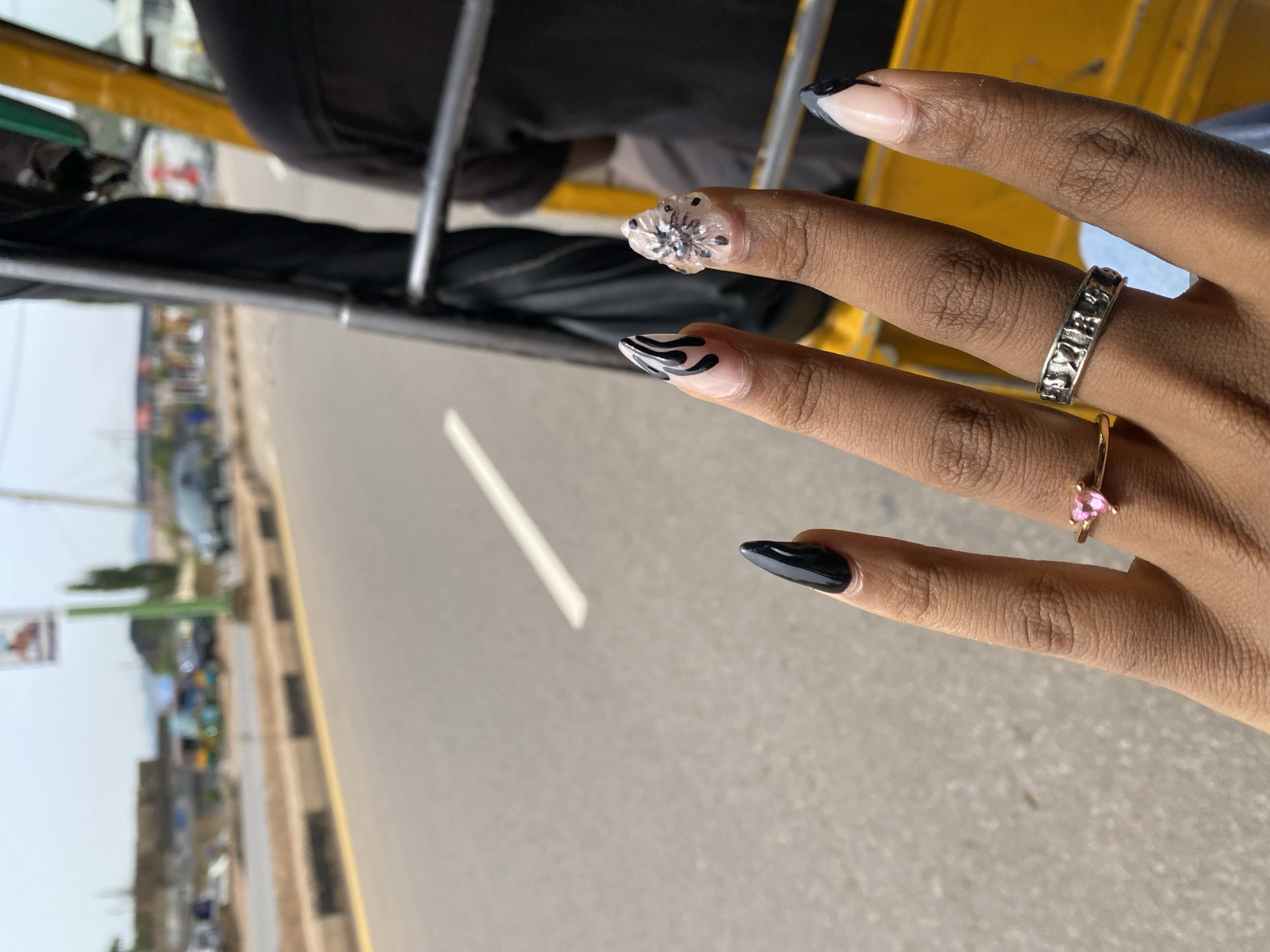 The image shows a close-up of a person's hand with manicured nails, resting on the metal frame of a vehicle, likely a tuk-tuk, given the context of Jos, Nigeria. The hand has long, pointed nails, some decorated with black and white abstract patterns, and one with a clear, three-dimensional flower design. The person is wearing two rings: a silver band with a textured pattern on the middle finger, and a delicate gold ring with a small pink heart-shaped stone on the ring finger.
The background is blurred, but it appears to be an outdoor scene. A paved road is visible, with a sidewalk and some vegetation along the side. In the distance, there are hints of buildings and what might be more vehicles. The lighting suggests it is daytime, possibly mid-morning or afternoon, under clear skies. The overall mood conveyed is casual and relaxed. No visible text is present in the image.