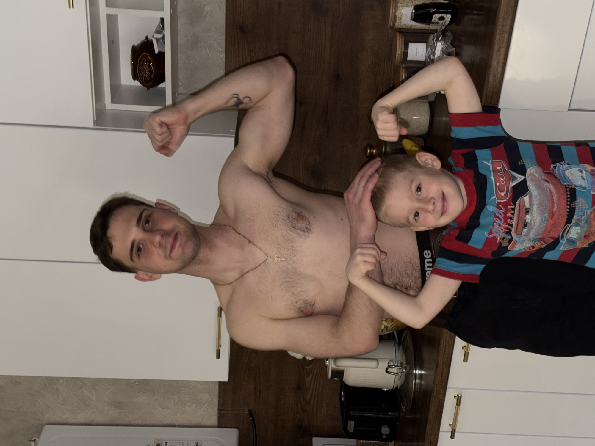 A shirtless man with short brown hair, wearing a gold chain, stands in a kitchen in Rossiyskiy, Russia, flexing his left arm. He has a tattoo on that arm. A young boy in a blue and red striped shirt with a Cars graphic is positioned next to him, also flexing his arm. The man's left hand rests on the boy's head. They are positioned on a dark wooden surface, likely a countertop, surrounded by kitchen appliances and storage. White cabinets are visible in the background, along with various containers and a decorative vase. A stainless steel kettle and black electronic device are on the counter. The lighting is bright.