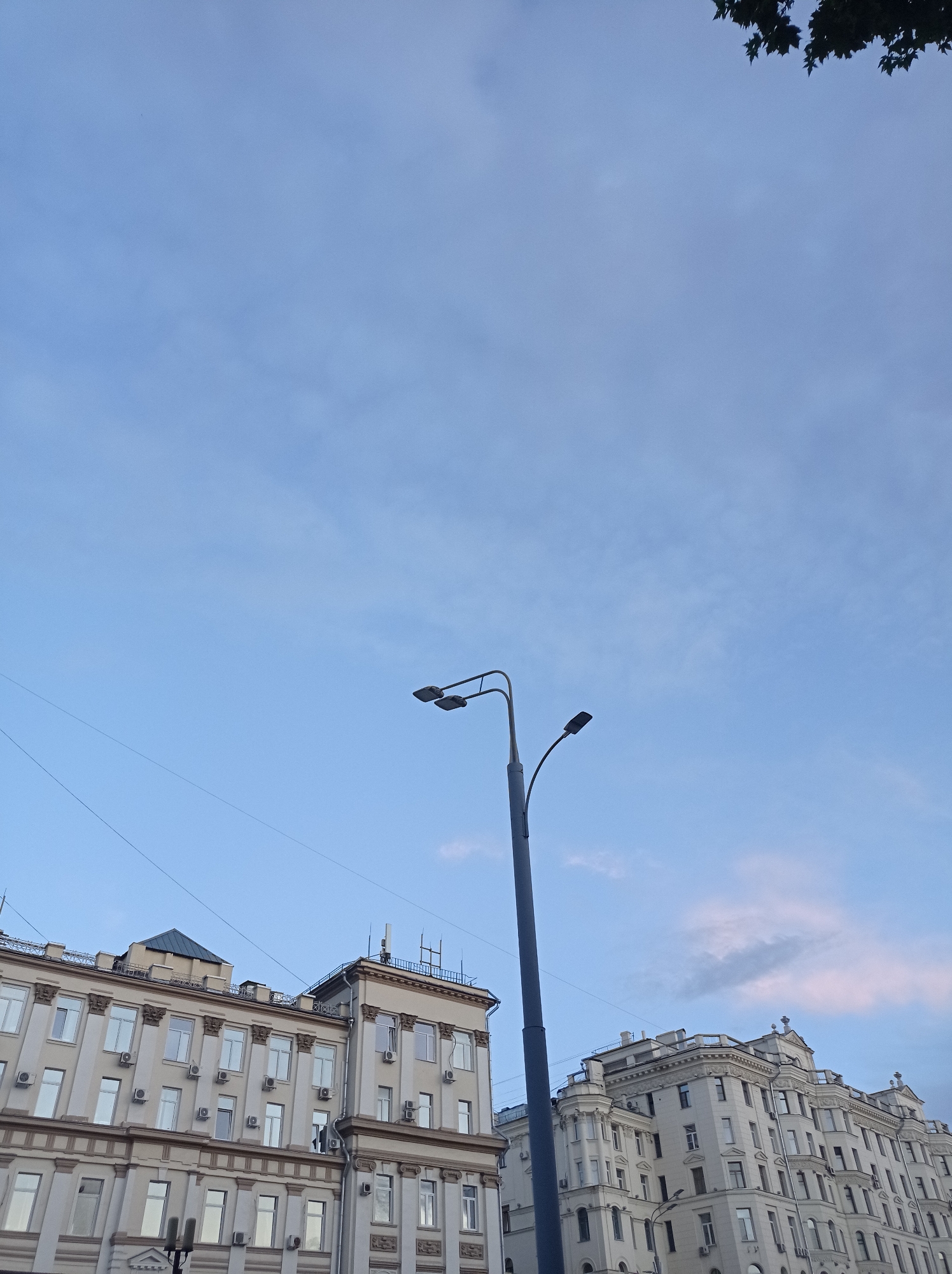 The image shows a street-level view of buildings and a lamppost in Moscow, Russia. The sky is a pale blue with some wispy clouds. The buildings are multi-story structures with numerous windows and ornate architectural details. There are several air conditioning units visible on the exterior of the buildings. A tall, dark lamppost with two lights extends upward into the sky. The buildings appear to be residential or commercial, with a style that suggests an older urban area. The overall impression is of a quiet, everyday scene in a Russian city.