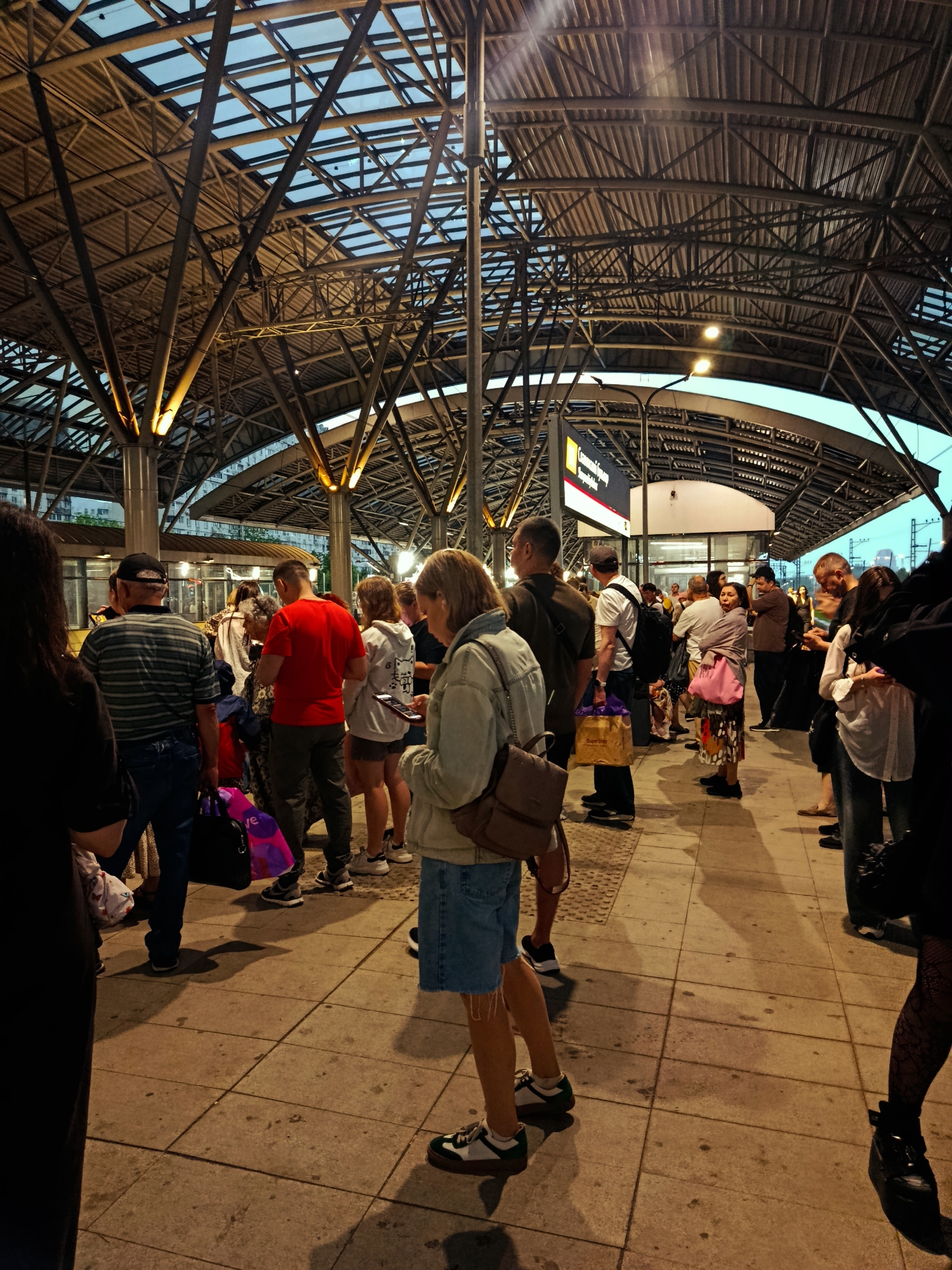 A crowd of people, appearing to be commuters, stand on a tiled platform under a large, modern, metal and glass canopy at a train station. The sky is visible through the transparent sections of the roof. The lighting suggests it is either early morning or late evening, with artificial lights illuminating the platform. The people are dressed in casual clothing and many are carrying bags, indicating they are either arriving or departing. In the background, train tracks and what appears to be a building with signage can be seen. The scene depicts a typical moment at a public transportation hub.
