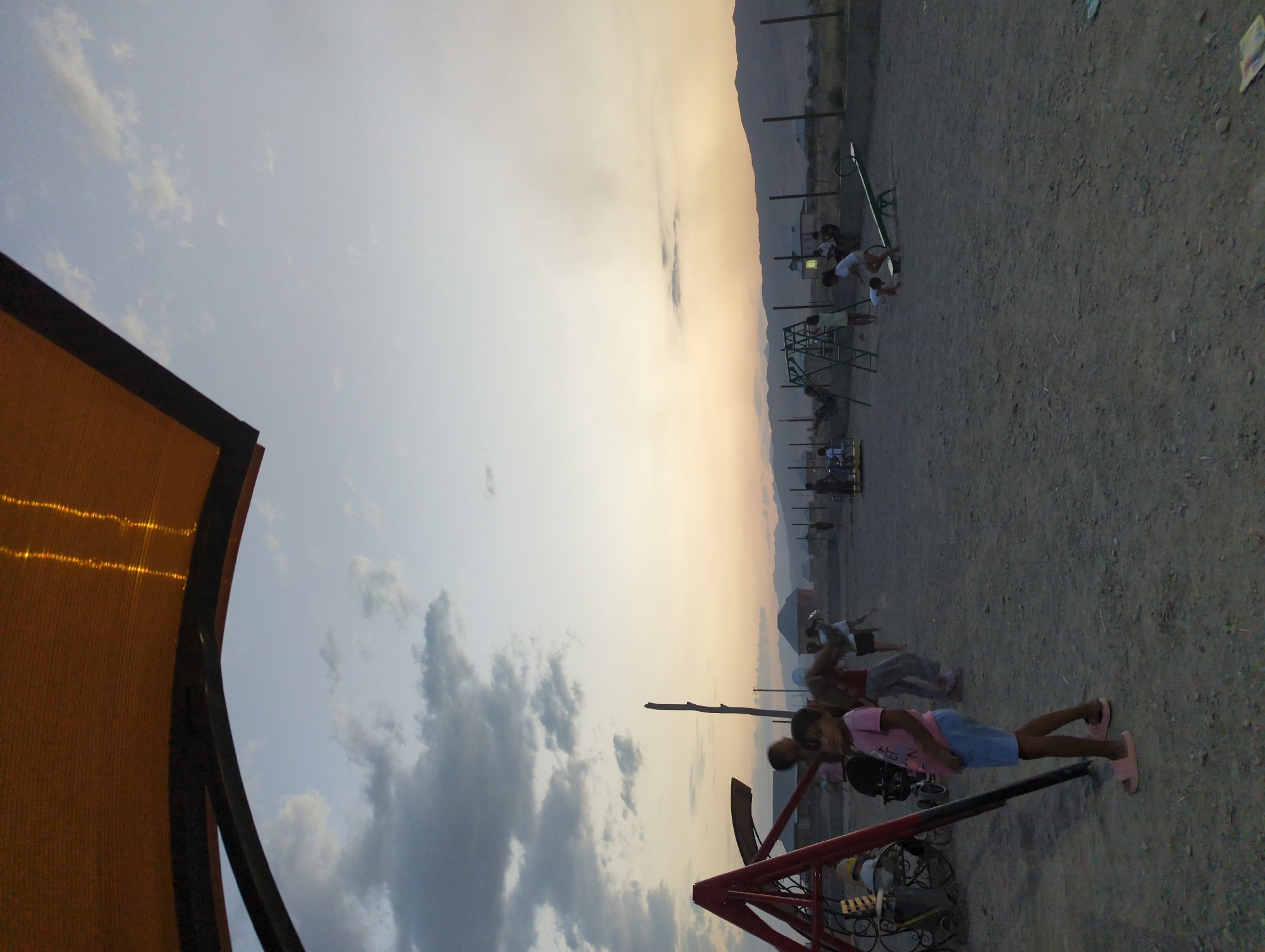 In an outdoor playground at dusk in the village of Üch-Döbö, Kyrgyzstan, several children are visible. A young girl in a pink t-shirt and blue shorts stands in the foreground, facing left. Behind her, another child is being swung on a red A-frame swing set. In the midground, other children are interacting with playground equipment, including a green jungle gym and a seesaw. The ground is covered in dirt and gravel. In the background, there are several low-rise buildings and a mountain range under a dusky sky. A large orange awning with visible lights extends from the top left corner of the frame.