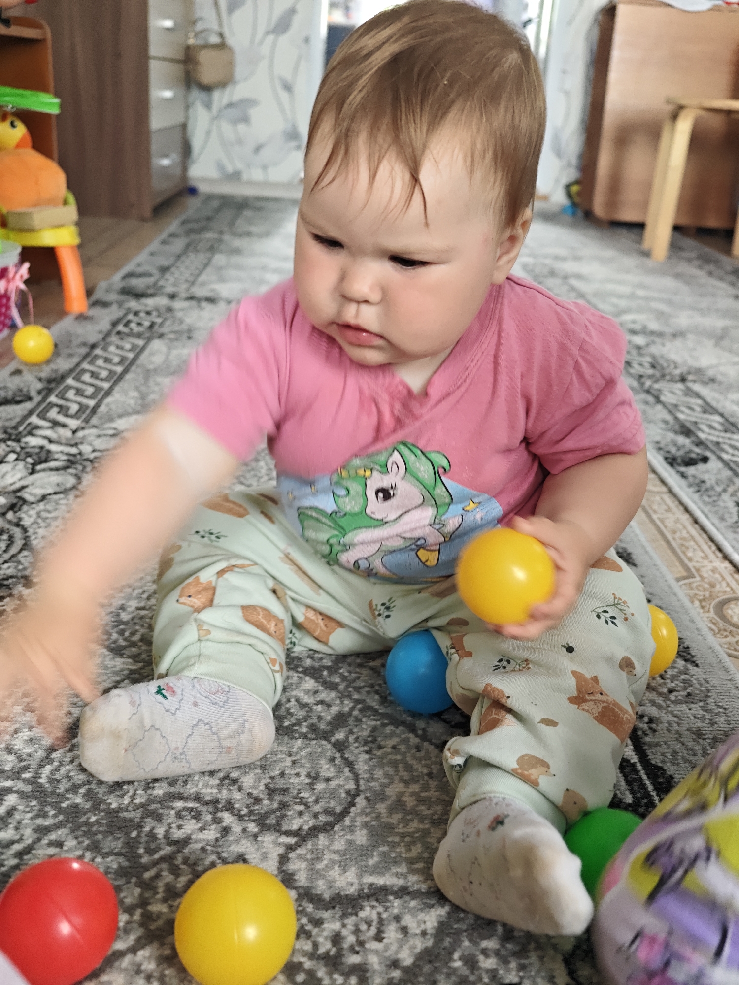 A baby sits cross-legged on a patterned grey rug indoors, surrounded by colorful plastic balls. The baby wears a pink t-shirt with a unicorn design and pale green pants with animal prints. The room includes a wooden dresser and a small stool in the background. The location is Уральское, Russia.