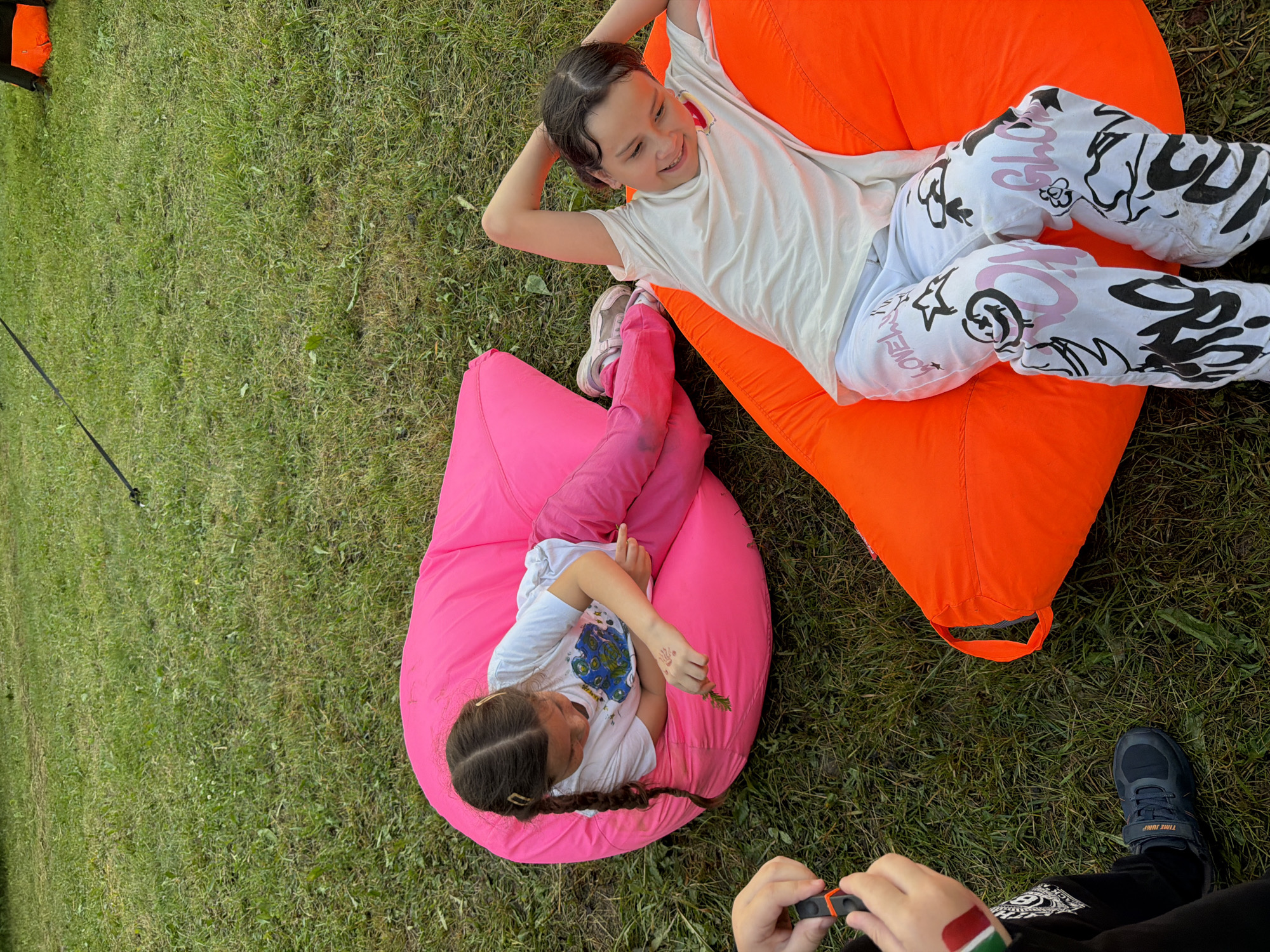 Two young girls are lying on beanbag chairs on a grassy area. The girl on the left is on a pink beanbag chair and is wearing a white t-shirt and pink pants. She is holding a small green plant and looking down at it. Her hair is in two braids. The girl on the right is on an orange beanbag chair and is wearing a white t-shirt and white pants with black graffiti-style drawings on them. She has her arms behind her head and is looking to her left with a smile. A third person's arm and hand are visible in the lower left corner of the image, holding a black object with orange accents. The scene appears to be outdoors in a park or backyard.