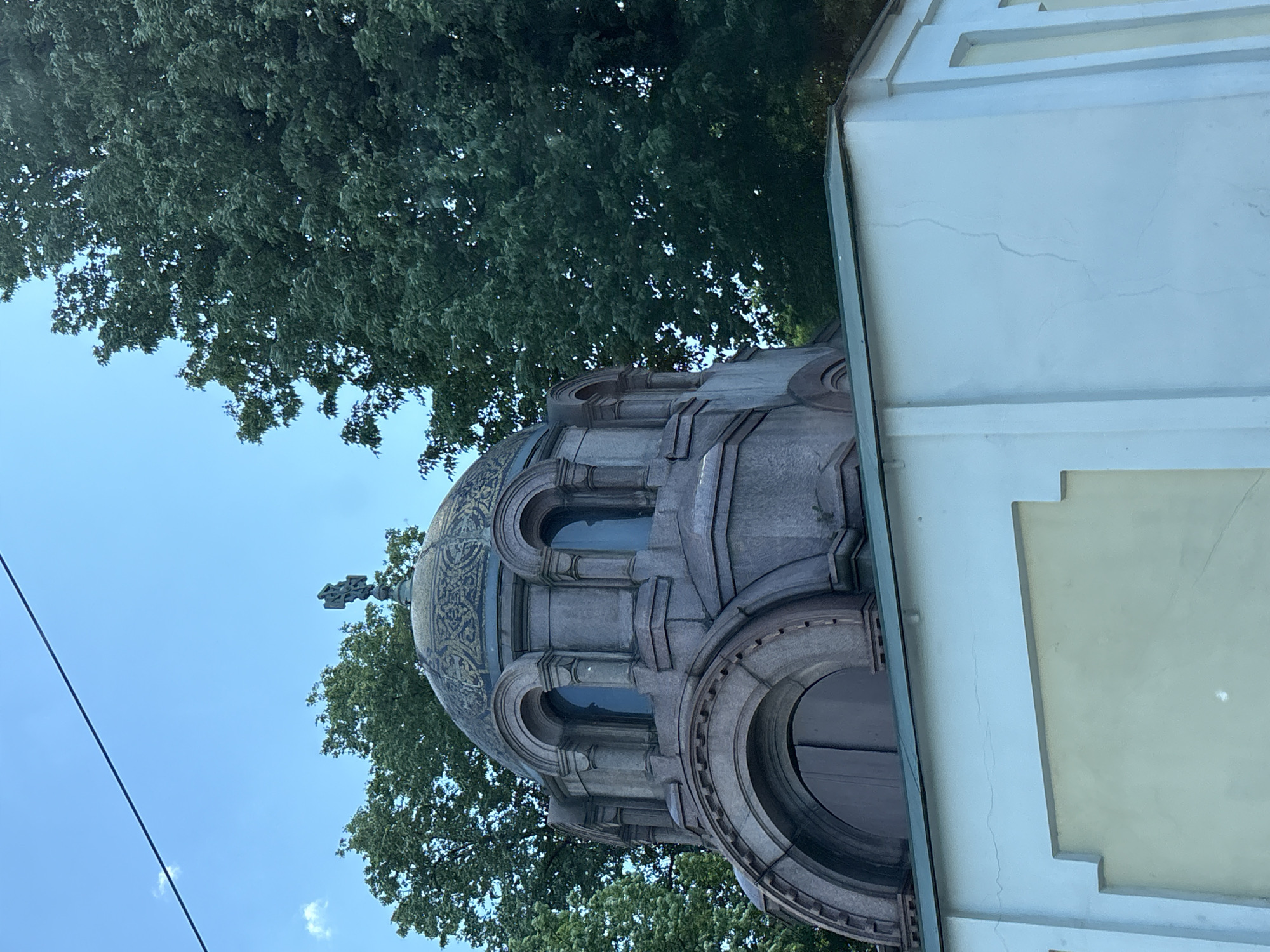 The image shows a close-up, low-angle view of the mausoleum of Count Fyodor Tolstoy, located in the Nikolskoye Cemetery of the Alexander Nevsky Lavra in Saint Petersburg, Russia. The mausoleum features a prominent, ornate dome with a cross on top, surrounded by arched windows and intricate stonework. The dome is adorned with what appears to be golden decorative patterns. The structure is made of stone, possibly granite, with a predominantly gray and pinkish hue. In the foreground, the white, modern-looking addition to the mausoleum complex is visible, with a pale yellow rectangular section. Lush green trees fill the background, and a bright blue sky with a few wispy clouds is overhead. A dark power line stretches diagonally across the upper portion of the sky. The lighting suggests it is daytime, likely late afternoon or early evening, given the soft shadows. There are no people visible in the image.