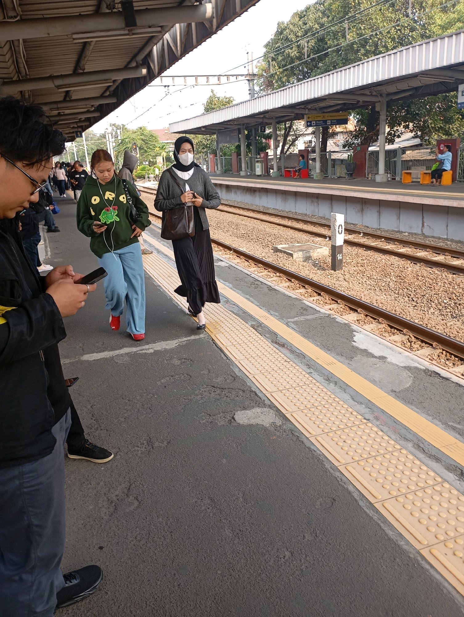 The image depicts a scene at a train station, likely in Tangerang, Indonesia, given the context of previous images from the same session. In the foreground, a person wearing glasses and a black jacket is looking at their phone. To their right, two women are walking on the platform. One woman wears a green hoodie with a frog graphic and is also looking at her phone. The other woman, wearing a gray cardigan and a black hijab, is carrying a brown bag. The background shows train tracks, a covered platform with supporting pillars, and trees. Several other people are visible in the background, waiting on the platform or walking. The platform has tactile paving for the visually impaired. A signpost with the number "0" and "14" is visible near the tracks. The overall environment suggests a busy public transportation hub.