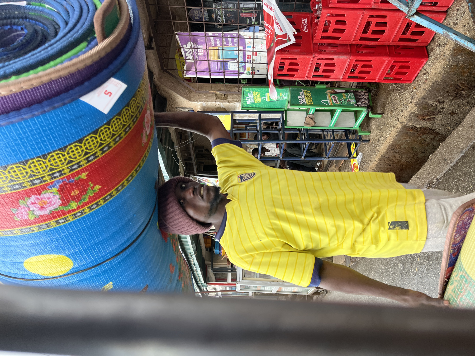 A man with dark skin, wearing a purple beanie and a yellow striped shirt with blue accents, stands in a street market or commercial area. He is positioned between a large roll of colorful mats on his left and possibly another stack of mats on his right. The scene includes red Coca-Cola crates stacked above him, and posters for "POS SERVICES AVAILABLE HERE", "PACK LUNCHBOX NOURISHING ENERGY" and Milo. Other objects include metal frames, and miscellaneous items. The location is identified as Jos, Nigeria based on the context. The man appears to be a vendor, possibly selling the mats.
